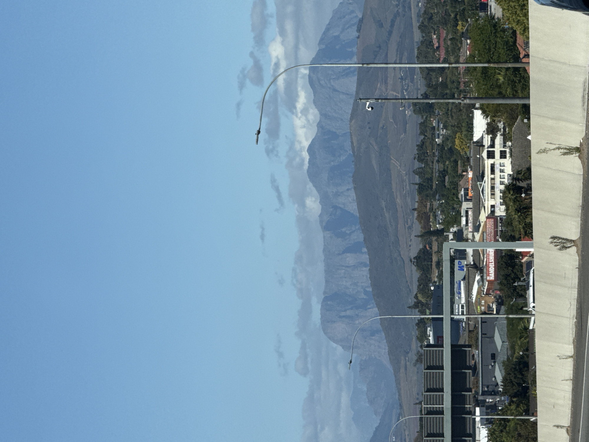 The image captures a view in Bellville, South Africa. In the background are layered mountains under a blue, slightly cloudy sky. A cityscape is situated along the base of the mountains. A grey concrete structure, possibly part of a road or bridge, is visible on the right side of the image. There are streetlights lining the road. Buildings, including commercial properties with signage like "HANDELSHUIS Auctioneers" are visible, interspersed with trees.