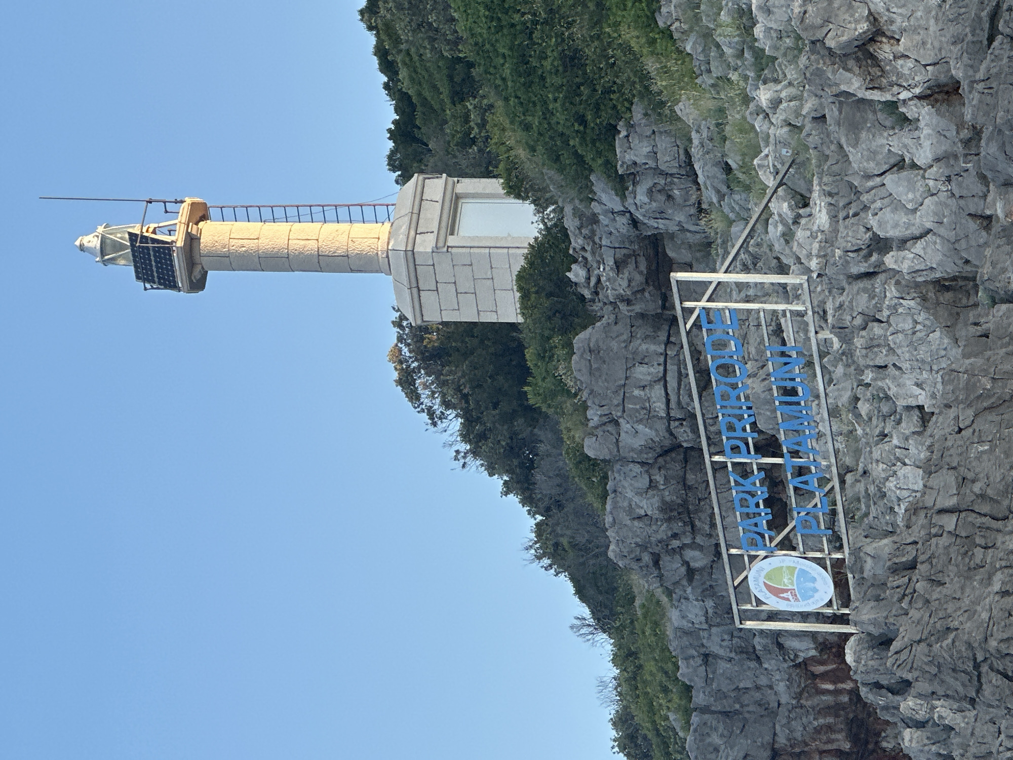 A stone lighthouse stands tall on a rocky, tree-covered hill overlooking the sea. In the foreground, a sign reads "PARK PRIRODE PLATAMUNI" with a circular logo containing a stylized depiction of a lighthouse and water. The sky is clear and blue. The scene appears to be located in Kubasi, Montenegro.