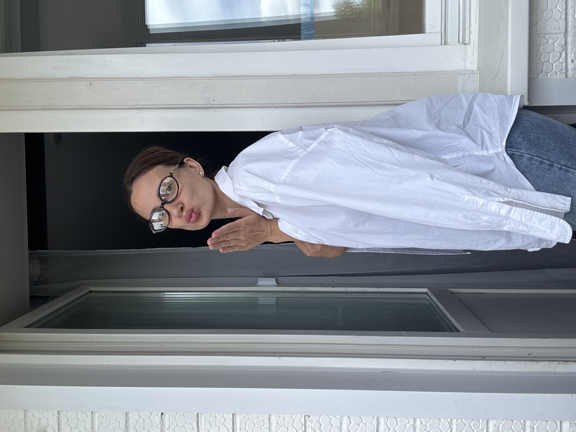 A young woman with brown hair and glasses is standing in a doorway, blowing a kiss. She is wearing a white button-down shirt and blue jeans. The doorway appears to be part of a building with white painted brickwork and a window. The background shows a curtain and a glimpse of the sky through the window. The woman is looking towards the camera. The scene appears to be outdoors, possibly on a balcony or porch. There is no specific indication of the location being in Chaikovsky, Russia.