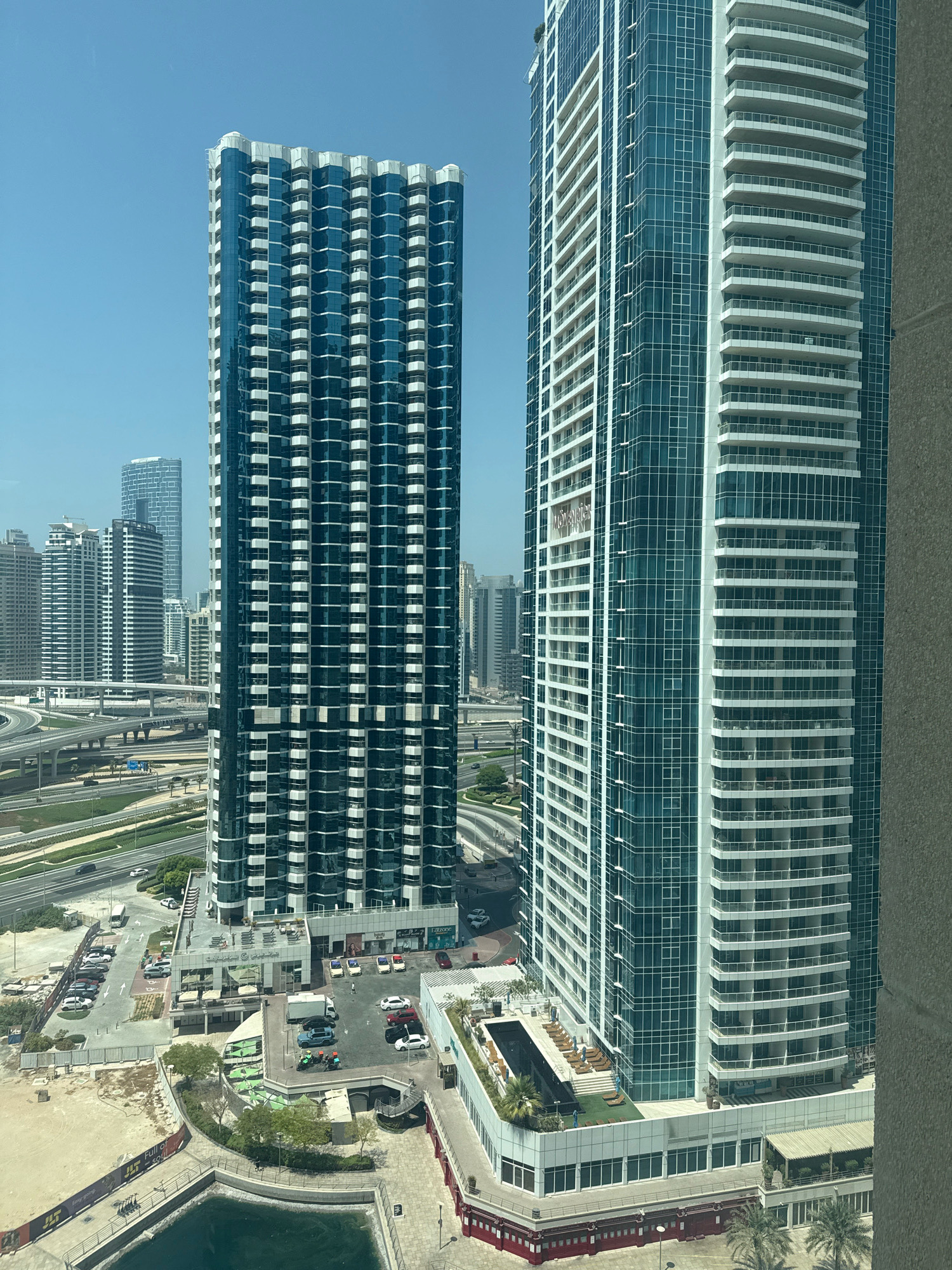 The image shows a view of the Jumeirah Lake Towers (JLT) district in Dubai, United Arab Emirates. Two prominent skyscrapers dominate the foreground: one on the left with a dark blue glass facade and white balconies, and a taller one on the right with a similar design but a lighter blue glass facade. Both buildings exhibit a repetitive pattern of floors with recessed balconies.
In the mid-ground, there is a complex network of multi-level highways and overpasses, characteristic of Dubai's infrastructure. Various vehicles, including cars and a white van, are visible on the roads and in parking lots. Adjacent to the skyscrapers, there are lower-rise buildings, including one with an outdoor seating area resembling a restaurant or cafe. Further in the background, other residential and commercial towers of varying heights and architectural styles are scattered across the cityscape.
The environment is bathed in bright sunlight, suggesting a clear, sunny day. The sky is a uniform, pale blue. At the bottom left of the image, a body of water, likely part of the JLT artificial lakes, is visible. The overall scene conveys a sense of modern urban development with a focus on high-rise living and efficient transportation infrastructure. No people are clearly identifiable in the image, nor are any specific actions or interactions taking place.