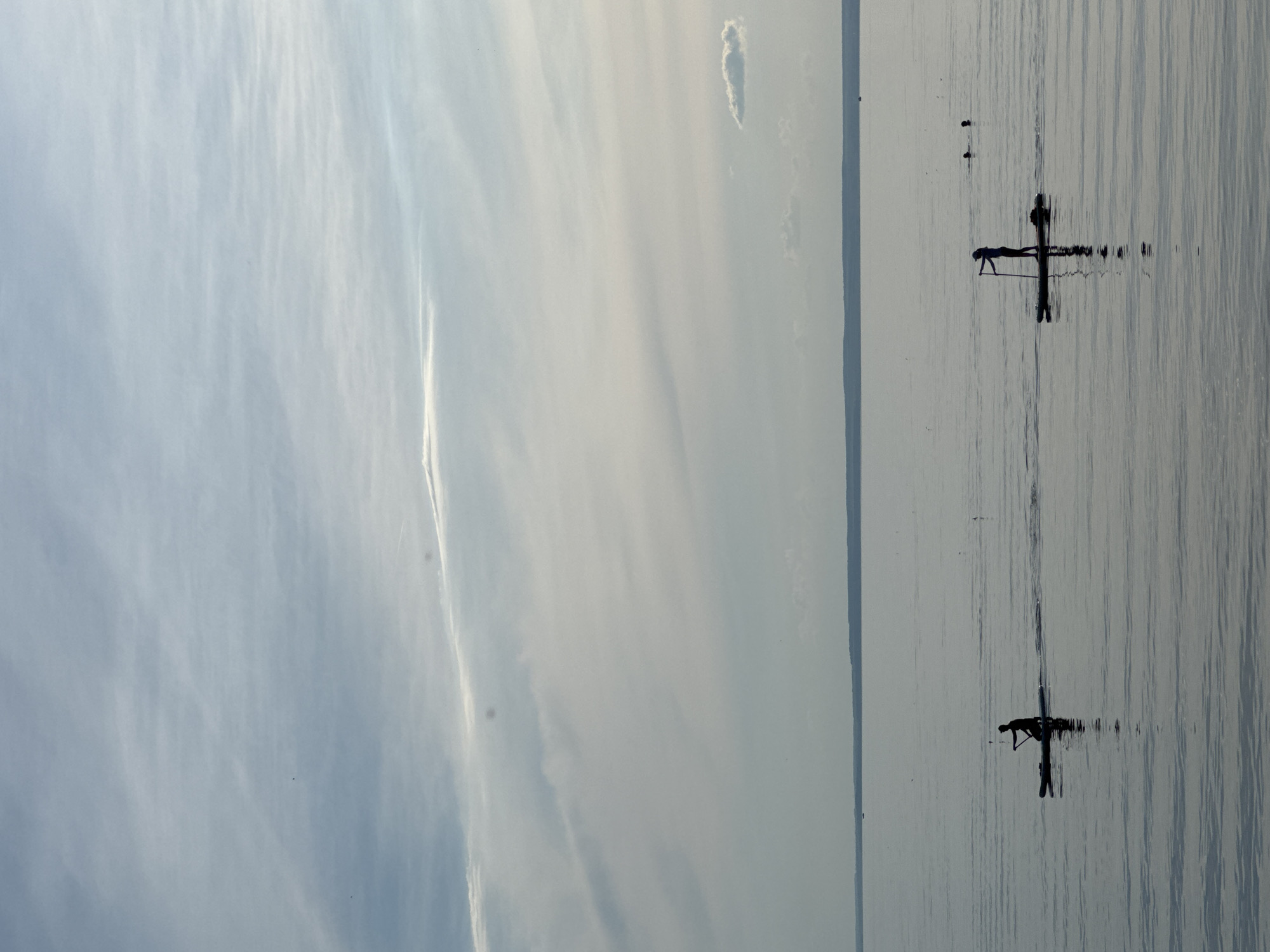 The media file shows a serene scene of a calm body of water, likely a lake or the sea, under a vast, softly clouded sky. The sky takes up the majority of the frame, exhibiting delicate, wispy clouds that create a sense of depth and movement. The water's surface is tranquil, marked by subtle ripples that reflect the light and the muted colors of the sky.
In the distance, silhouetted figures can be seen on the water. The most prominent subject is a person on what appears to be a paddleboard or a similar watercraft, striking a pose that suggests an activity like yoga or meditation. The individual's silhouette is sharp against the reflective water, creating a striking contrast. The pose is an inverted V shape, with one leg extended upwards and the arms supporting the body. The reflection of this figure is clearly visible, mirroring the person and their watercraft on the calm surface. Another smaller, distant silhouetted figure is visible on the water, slightly further away, suggesting there might be more than one person engaged in activities on the water.
The lighting suggests it is either early morning or late afternoon, possibly during the "golden hour," due to the soft, diffused light and the gentle colors in the sky. The overall atmosphere is one of peace, tranquility, and solitude. The vastness of the sky and the stillness of the water contribute to a feeling of calm and introspection. There is no visible text in the media file. The setting is natural and open, devoid of any man-made structures or prominent landmarks, emphasizing the connection between the individual and the natural elements.