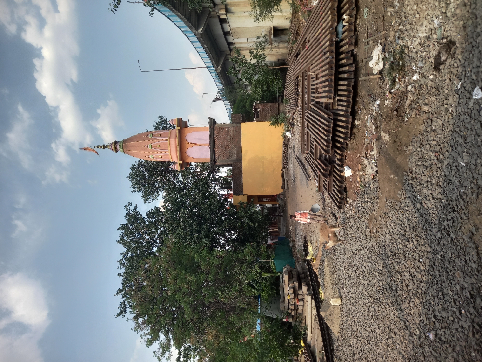 The image depicts a Hindu temple in Bhopal, India, situated near construction activities. The temple has a distinctive dome and is painted in shades of orange and yellow. It stands next to a brick building with a yellow wall. In the foreground, there's a pile of grey gravel and discarded materials, suggesting construction or renovation work. Metal beams are stacked nearby. A man in a red and white shirt is walking in the scene, carrying a construction tool. A light brown dog is standing next to him. The backdrop includes a bridge and lush green trees, with a partially cloudy sky.