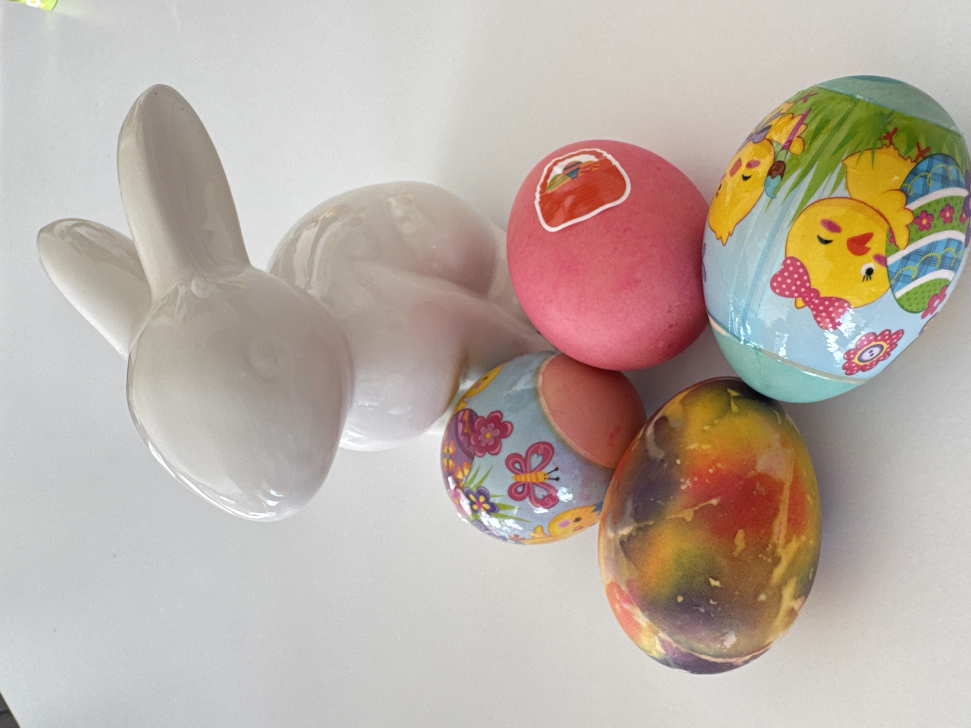 A high-angle shot depicts a white ceramic rabbit figurine next to four painted Easter eggs on a white surface. The eggs vary in color and design. One is pink with a sticker, another features cartoon chicks on a light blue background, a third is light blue with butterfly and floral designs, and the last egg has a multi-colored, marbled pattern. The location is unspecified and has no relation to Varna, Bulgaria.