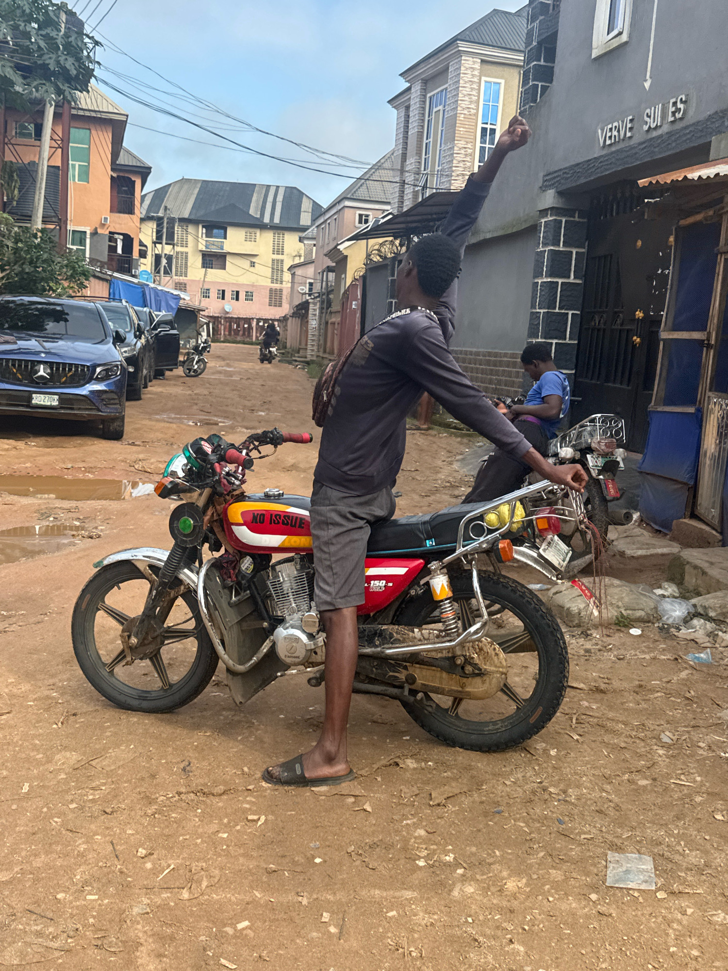In Ihiagwa, Nigeria, a young Black man stands beside a red and yellow motorcycle in a dirt road. He wears a dark long-sleeved shirt and grey shorts, with his right arm raised. The motorcycle has "NO ISSUE" written on its side. In the background, other motorcycles and blue vehicles are parked. Buildings line the street, with one featuring the sign "VERVE SUITES". The sky is overcast.