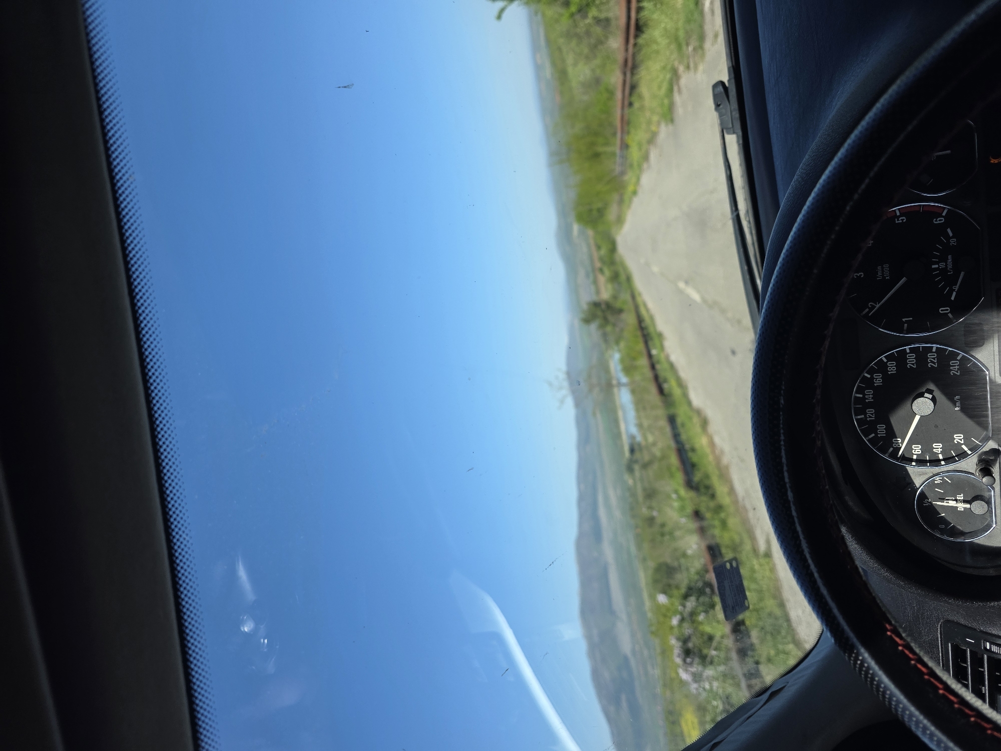 The image captures a first-person perspective from inside a car, looking out through the windshield onto a scenic rural landscape in Lelintsi, Bulgaria. The vibrant blue sky, mostly clear with a few dust specks on the glass, dominates the upper part of the frame, indicating a bright, sunny daytime. A paved road stretches into the distance, appearing to wind through rolling green hills and valleys. Lush green vegetation, including what might be trees and grass, lines the roadside and covers the distant terrain. A body of water, possibly a river or lake, is visible in the middle distance.
Inside the car, the dashboard and part of the steering wheel are visible in the foreground, primarily on the right side of the image. The steering wheel is black, textured, and features red stitching. The instrument cluster on the dashboard includes a speedometer displaying a reading of approximately 60 km/h, a tachometer showing 0 x1000 1/min (0 rpm), and a fuel gauge indicating a fuel level slightly below half, with the word "DIESEL" clearly visible. An orange warning light is illuminated on the dashboard. The windshield shows reflections and some minor dust or streaks, along with the black frit band on its left edge. The scene suggests a journey in progress or a stop along a picturesque route.