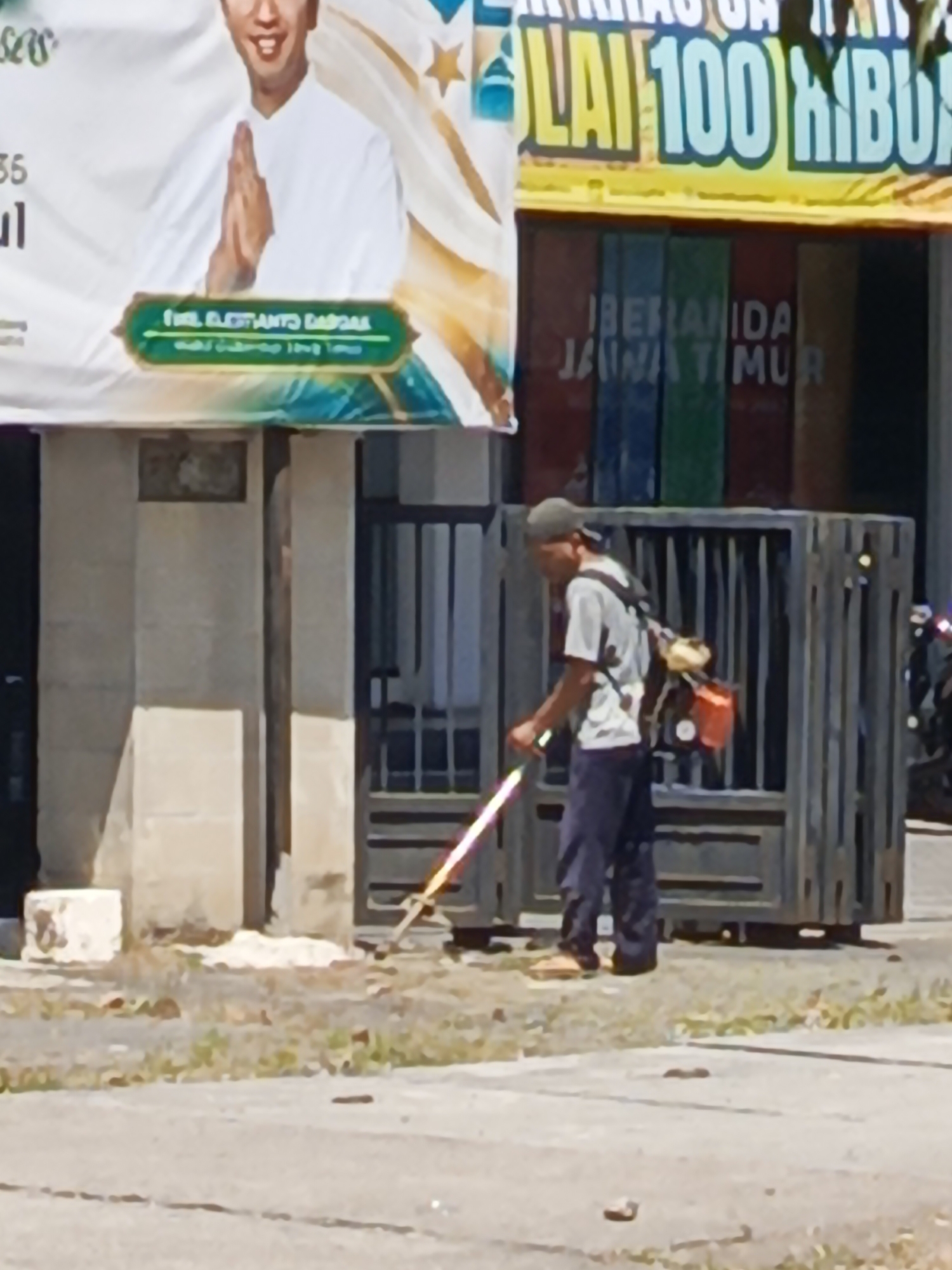 A man wearing a grey t-shirt, dark pants, and a baseball cap is operating a weed trimmer. He has a backpack power source attached to his back. The trimmer has a long shaft and a blade at the end which appears to be emitting a reddish-orange light. The man is positioned in front of a metal gate with vertical bars and a wider horizontal bar at the top. Behind the gate, there are several tall, vertical colored panels, appearing to be red, blue, and green. To the left of the gate, there is a light-colored, rough-textured wall. Above the wall and gate, a large banner is visible. The banner features a picture of a man in a white shirt with his hands pressed together in a greeting gesture, and text in green and yellow. Further text is visible to the right of the banner, with large yellow lettering on a dark background, and white text layered on top. The ground in front of the gate is a mixture of dirt and sparse green vegetation. To the far right, a motorcycle is partially visible. The scene is outdoors, likely in a public area.