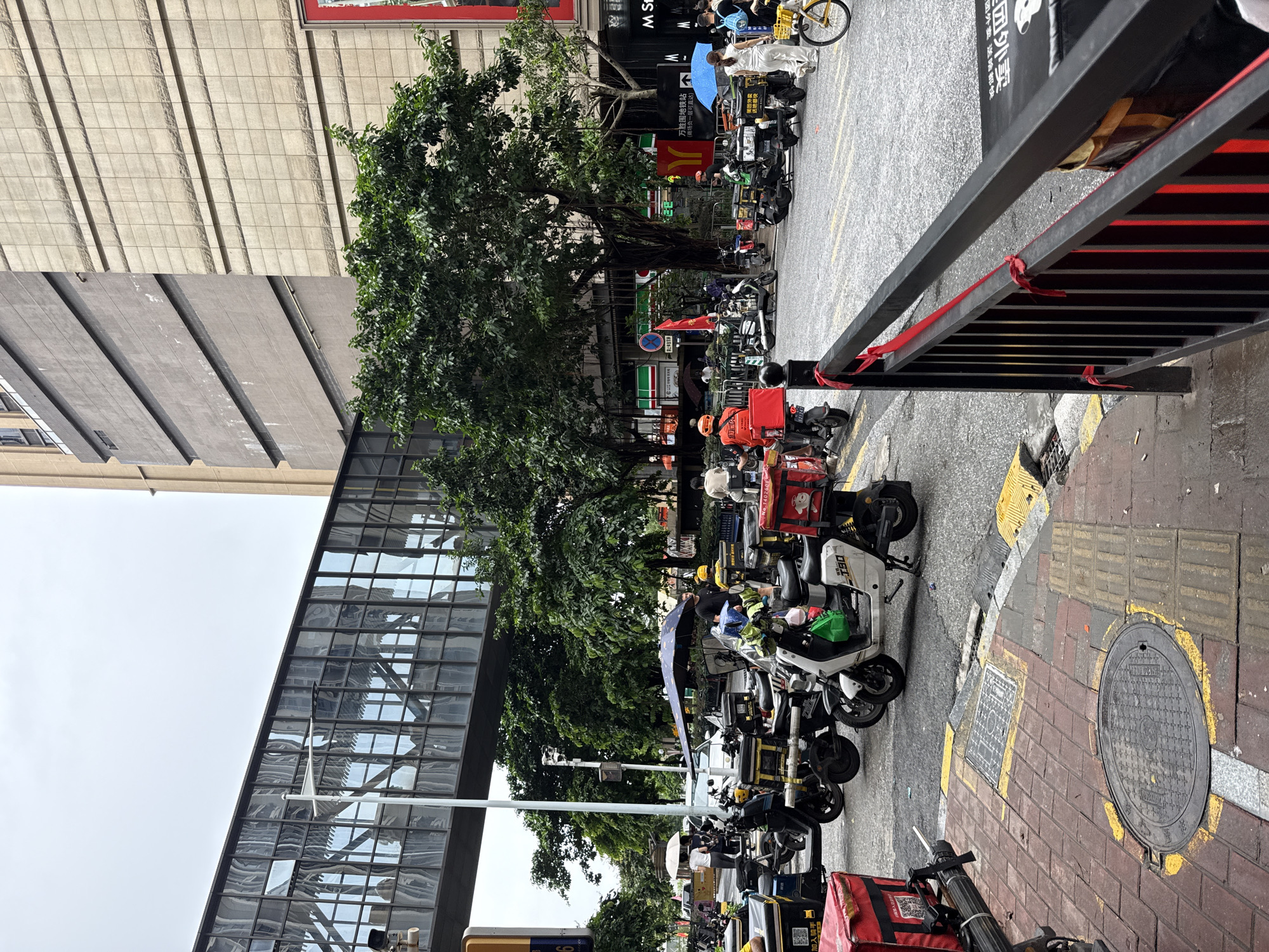 This image depicts a street scene in Haizhu District, China, characterized by a skywalk connecting two buildings, numerous delivery motorcycles, and pedestrians. The skywalk, made of glass and metal, spans across the street at a high level, reflecting the surrounding environment. Below, a fleet of delivery motorcycles, many equipped with large food delivery boxes, are parked along the roadside. Several delivery personnel, identifiable by their uniforms and helmets, are either attending to their vehicles or waiting. Pedestrians are also visible, some walking along the sidewalk and others crossing the street, with a few holding umbrellas due to overcast conditions. The environment includes trees lining the street, a large building with a grid-like facade, and various street furniture such as traffic signals and fences. The presence of delivery vehicles and personnel suggests an active urban area, likely a commercial or residential district.