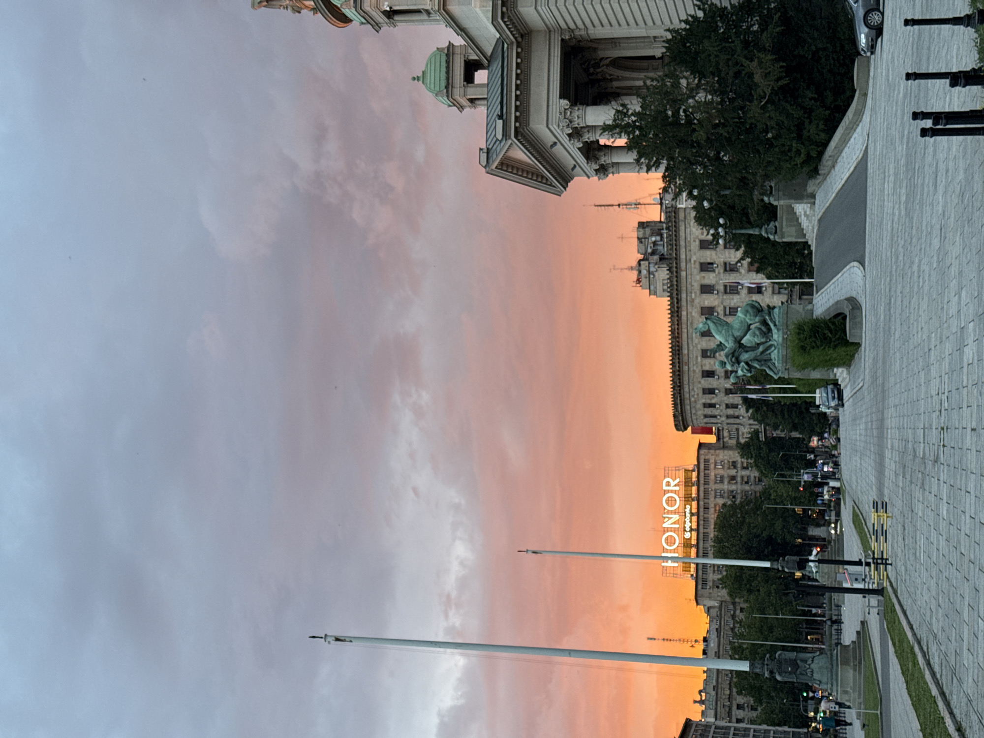 The image captures a sunset over Belgrade, Serbia, with a view of the National Assembly building and the surrounding area. The sky is filled with vibrant orange and pink hues, indicative of a dramatic sunset. In the foreground, a paved walkway leads towards a grand, classical-style building with columns and a green dome, which is the National Assembly. To the left of the walkway, two tall flagpoles stand prominently. Across the street, a large building features a bright "HONOR" sign illuminated against the darkening sky, along with a smaller sign for "otpbanka". A bronze equestrian statue is visible on the right side of the walkway, adding a touch of artistry to the scene. Several trees are scattered throughout the area, and a few cars are parked or driving in the background. The overall impression is one of urban tranquility and natural beauty, with the city's architecture serving as a backdrop to the breathtaking sky.