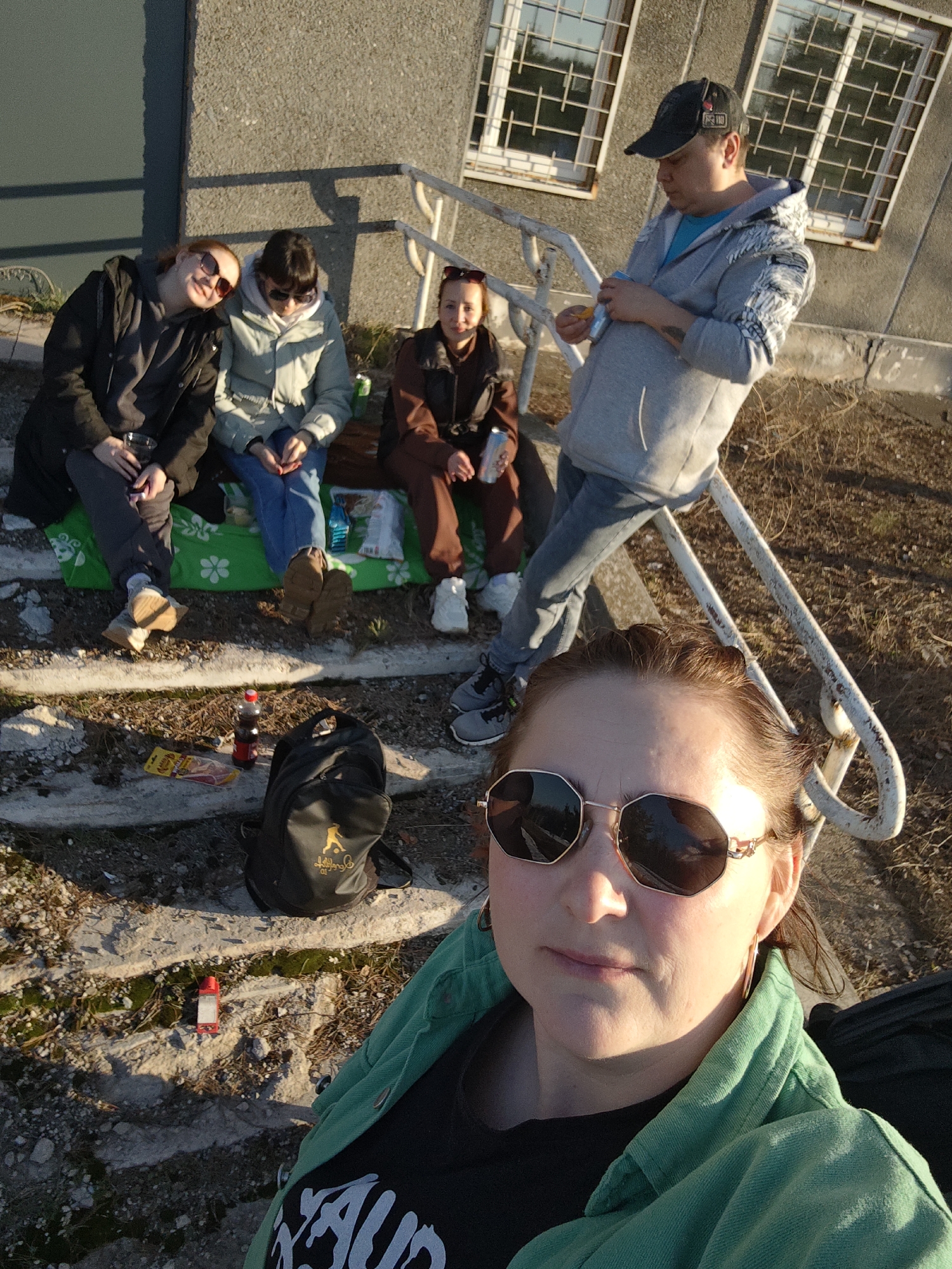 A group of five individuals are gathered on and around a set of concrete steps near a building in Марковский, Russia. Four people are seated on the steps, utilizing a green and white patterned blanket, while one individual stands to the side, leaning against a white railing. The foreground features a woman taking a selfie, wearing sunglasses and a green jacket. The setting appears to be outdoors, likely in the late afternoon or early evening, as indicated by the long shadows and warm lighting. Objects visible include bottles, a snack package, and a black backpack on the steps. The building behind them has barred windows and a grey facade. The overall atmosphere suggests a casual social gathering.