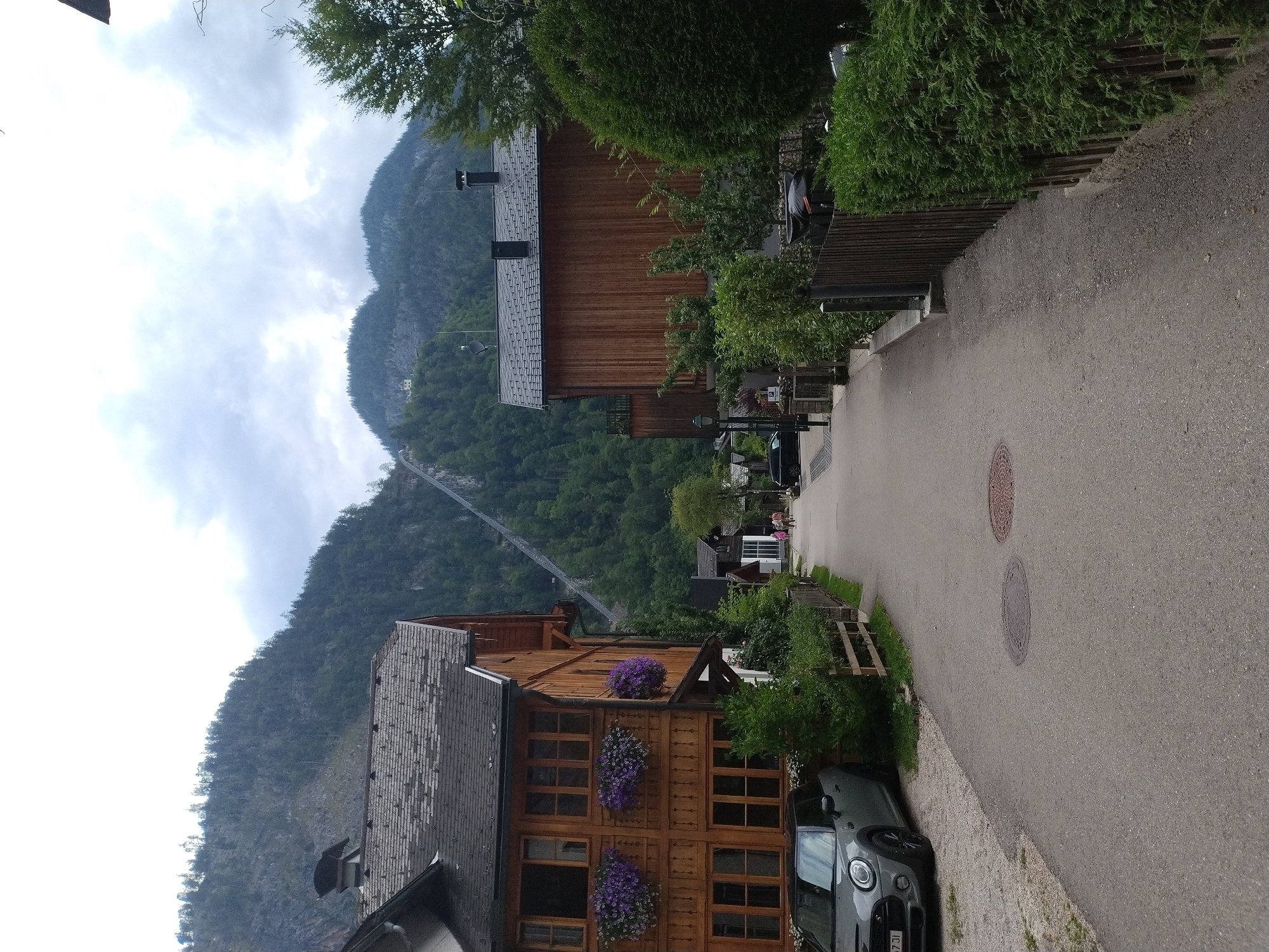 The image shows a street scene in Hallstatt, Austria. On the left, a two-story wooden building with a dark shingled roof is visible. It has several windows with planters filled with purple flowers. A grey Mini Cooper car is parked in front of the building. A wooden fence separates the property from the street. On the right side of the street, another wooden building with a dark shingled roof is seen, along with a fence and dense green hedges. A paved road runs through the center of the scene, with manhole covers on the surface. In the background, a steep, tree-covered mountain rises, with a long, thin structure, possibly a funicular railway or a zipline, ascending its slope. The sky is overcast with clouds. A few people are walking down the street in the distance. The overall impression is a quaint, European village setting nestled against a mountainous backdrop.
