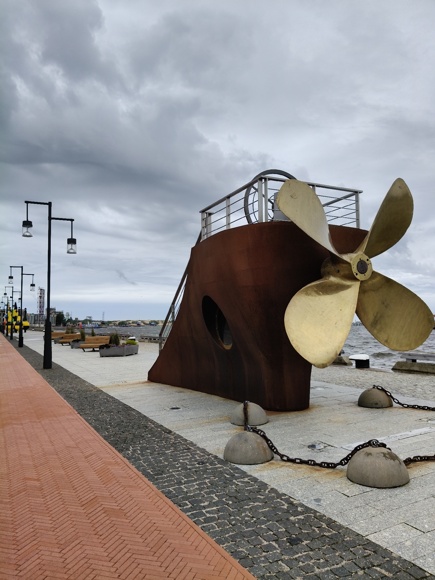 The image depicts a promenade along the waterfront in Ventspils, Latvia. A large, weathered metal sculpture resembling the stern of a ship, complete with a prominent brass propeller, dominates the foreground. The sculpture is secured to the ground with heavy chains attached to concrete bollards. To the left, a brick pathway with a herringbone pattern runs parallel to a walkway paved with grey cobblestones. Several black lampposts with a modern design are spaced along the walkway. Further back, there are benches, planters, and in the distance, more of the city's waterfront can be seen, including industrial structures and buildings. The sky is overcast with heavy grey clouds, suggesting a cool or potentially rainy day. The sea is visible on the right side of the image, with waves indicating a moderate breeze. No people are clearly visible or engaged in any specific actions within the frame.