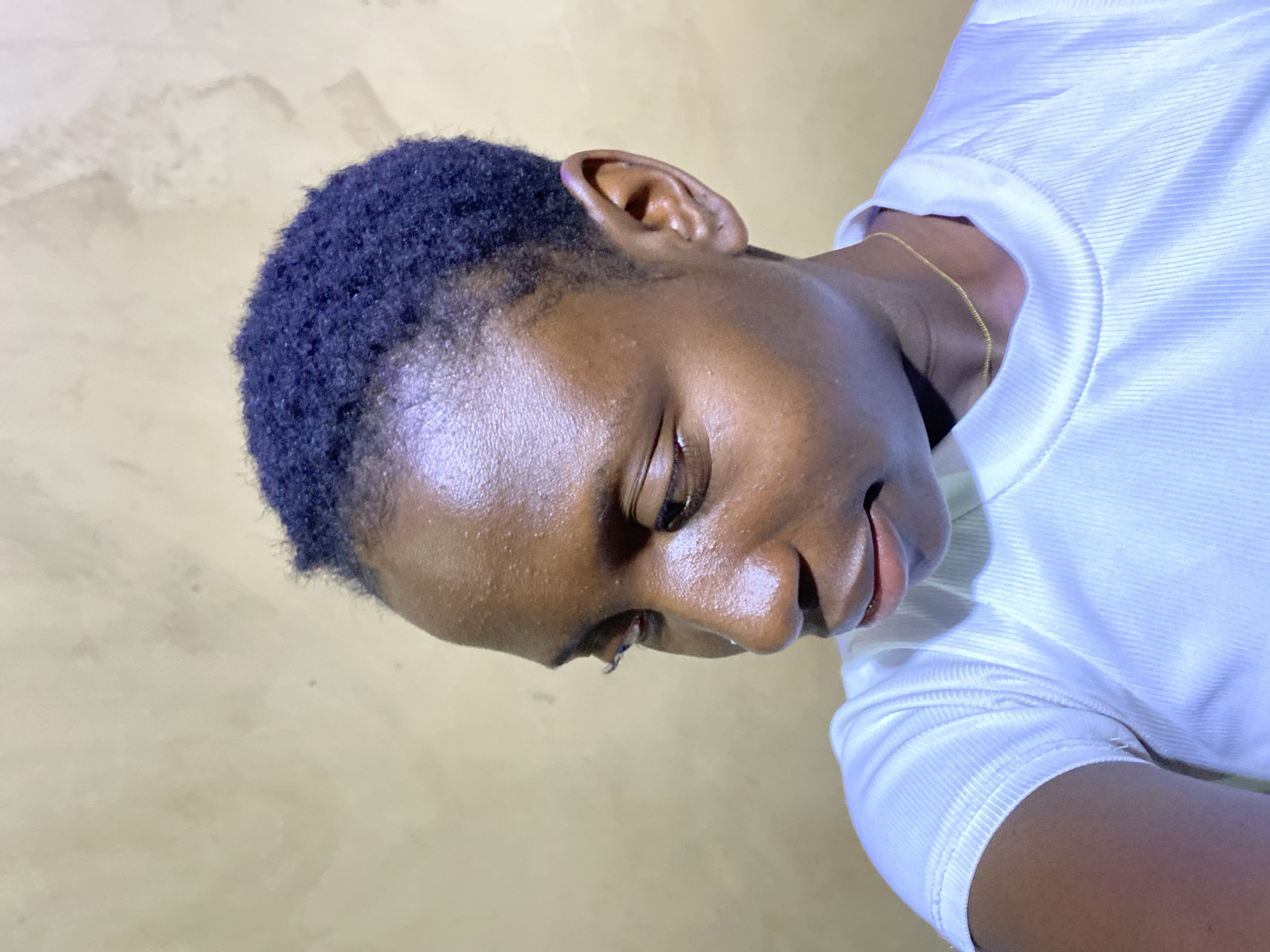 A portrait-style image, rotated 90 degrees counter-clockwise, features a person with dark skin and short, dark, tightly curled hair. The subject's head is angled downwards and to the side, with their gaze directed off-frame, suggesting a pensive or relaxed mood. Their lips show a very subtle, gentle curve, hinting at a slight smile. The skin on their forehead, cheek, and nose appears somewhat moist or shiny, possibly due to sweat or natural oils.
The person is wearing a plain white t-shirt or top with a distinct ribbed texture, visible clearly on the sleeves and collar area. A thin, delicate gold chain is worn around their neck, resting just above the collarbone. Their right ear is fully visible, showing its natural folds.
The background is a simple, light beige or sandy-colored wall with slight textural variations and subtle, darker markings that suggest a painted or rendered surface. The lighting is soft, highlighting contours and the sheen on the skin. No specific time of day or weather can be definitively determined, nor is any text visible. The image captures a quiet moment, likely taken indoors or against a simple wall in Kuru Dungus, Nigeria.