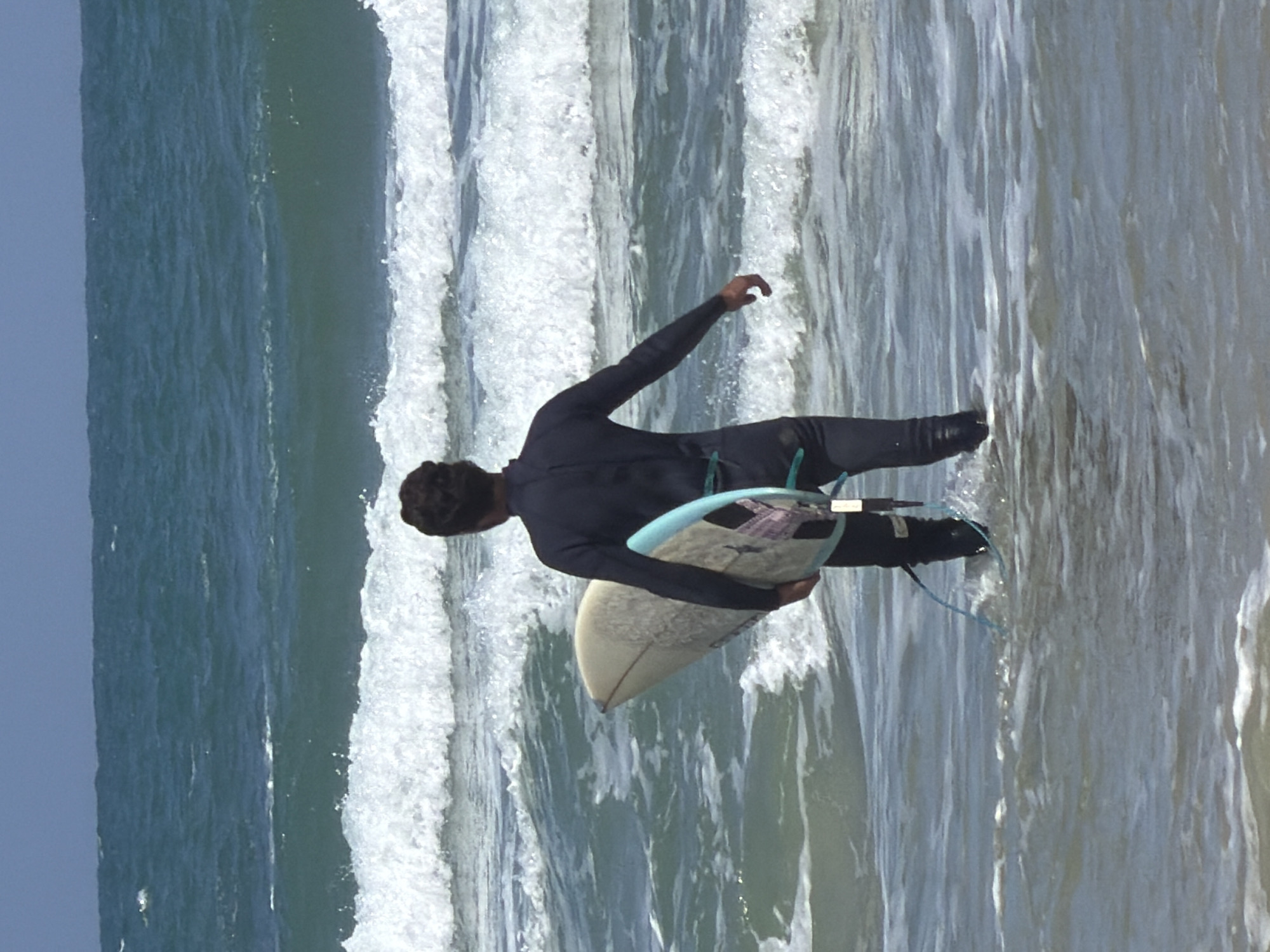 A surfer dressed in a black wetsuit is standing in the water with a surfboard under their arm. The water is choppy with white-capped waves. The sky is overcast. The scene is a beach, likely located in Tel-Aviv, Israel, considering the context.