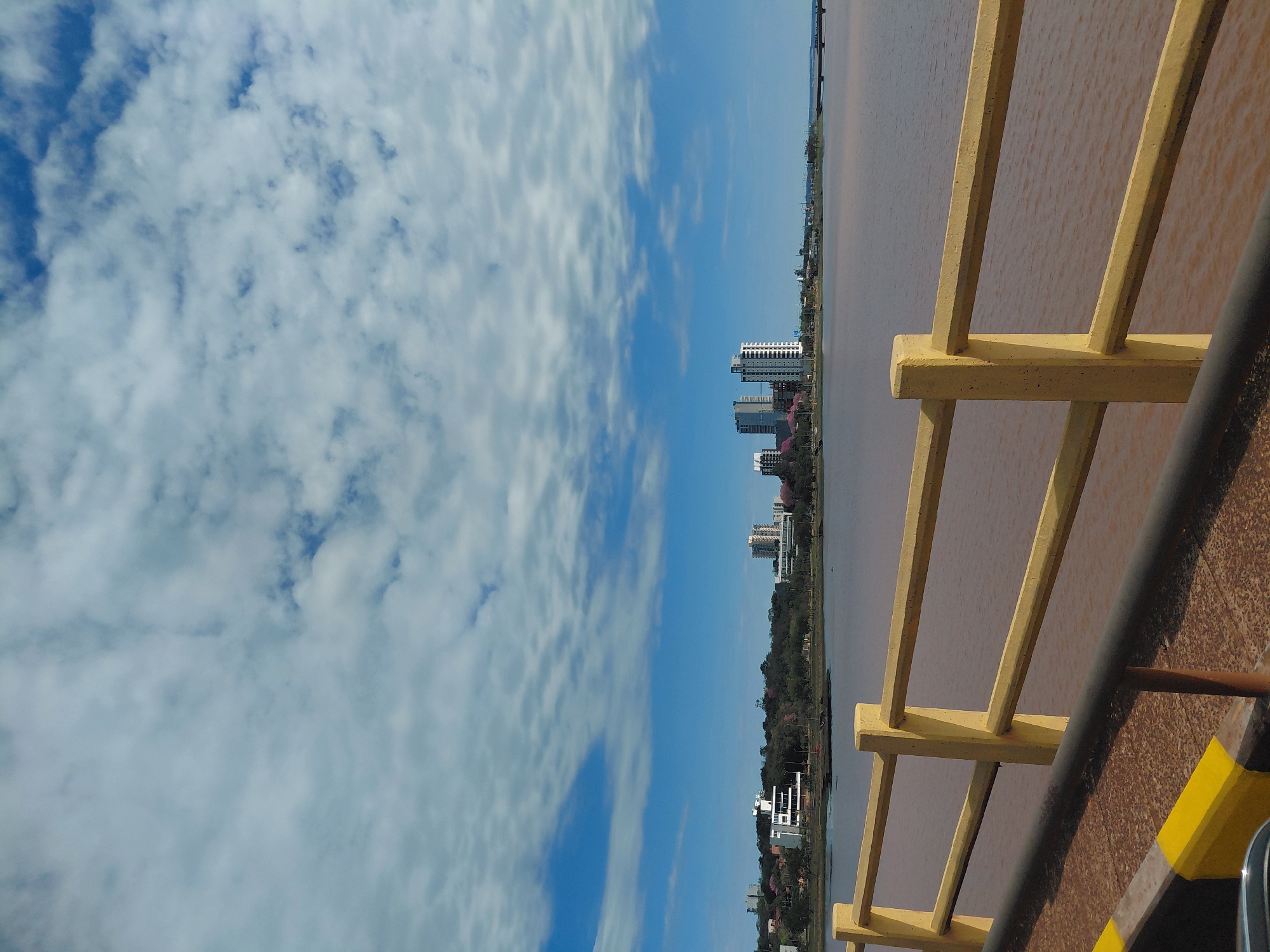 The image displays a scenic view from a bridge overlooking a body of water with a city skyline in the distance. The foreground features a yellow railing with a gray metal bar below it, indicating a walkway or bridge structure. The water appears to be a reddish-brown color, possibly due to sediment or algae.
In the midground, the city of Encarnación, Paraguay, is visible with several modern buildings and a tree-lined shore. Notably, there are a few high-rise buildings, suggesting a developed urban area. The sky is a vibrant blue with scattered white clouds, creating a dynamic and somewhat overcast atmosphere. The presence of a bridge structure in the foreground suggests a viewpoint that is elevated, offering a panoramic perspective of the city and the waterway. The overall scene depicts a tranquil day with a blend of urban development and natural landscape.