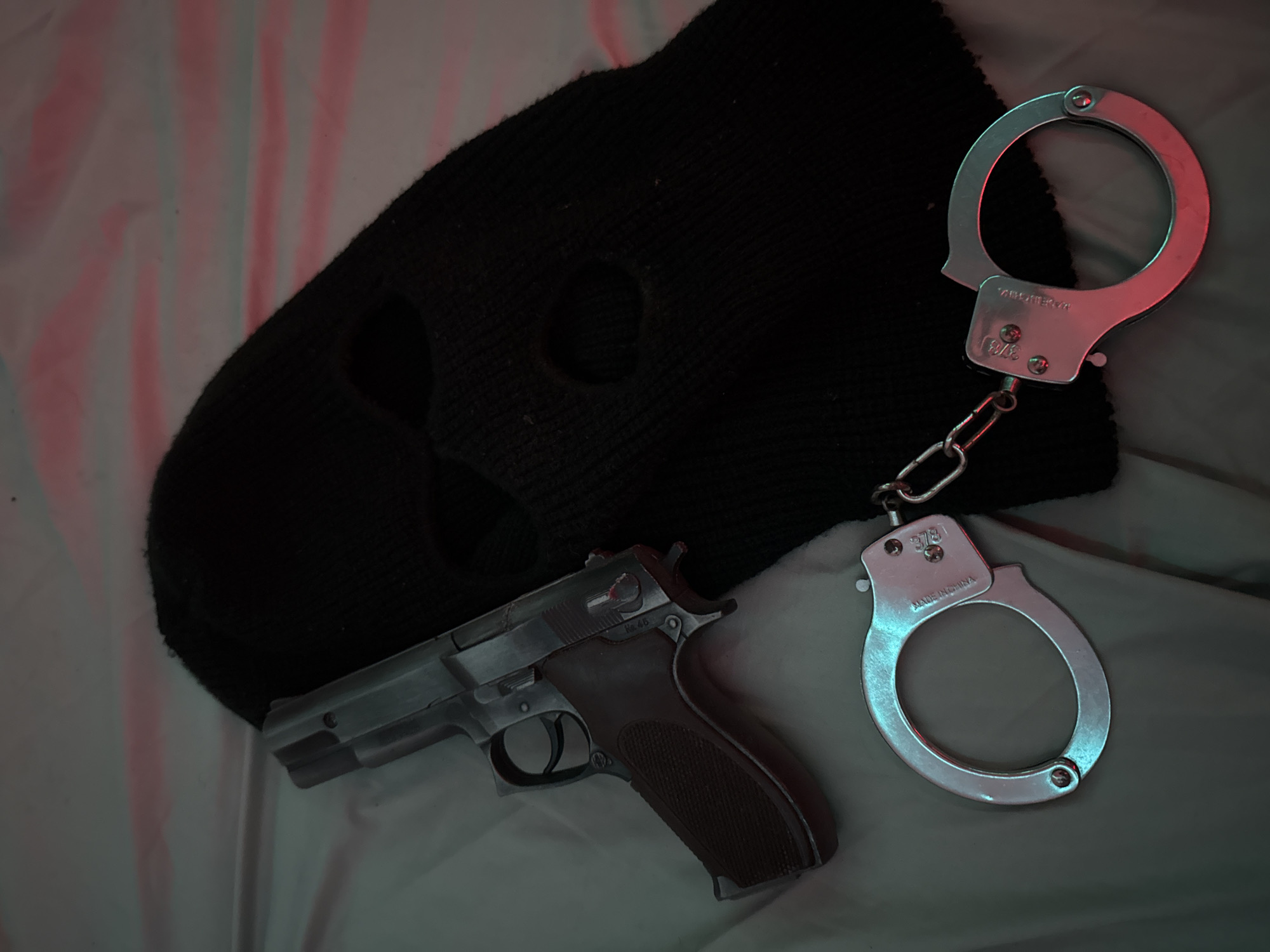 A balaclava, a handgun, and a pair of handcuffs are arranged on a white sheet with pink and blue lighting. The balaclava is black and has eye and mouth holes. The handgun is metallic with a brown grip. The handcuffs are silver and appear to be made of metal. The scene is dimly lit, suggesting it may be nighttime or indoors. There is no indication of the specific location within Varna, Bulgaria.