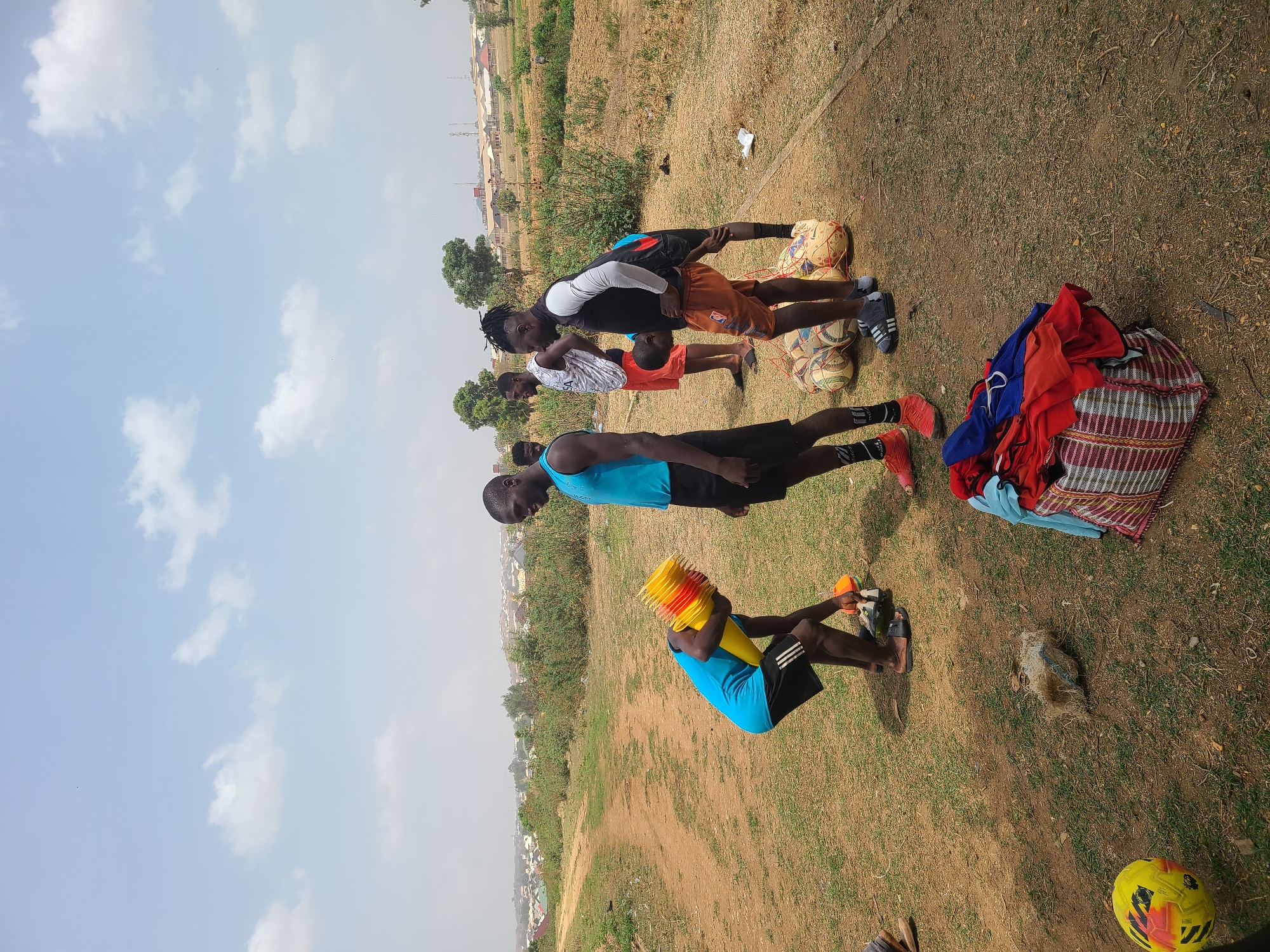 The image depicts a group of young men outdoors in Barikin Jirga Summa, Nigeria, likely on a field or training ground. Four men are visible, each engaged in or related to physical activity. One man is seated on the ground, possibly resting, holding a stack of yellow and orange plastic cones. Another man stands with a blue tank top and black shorts. Two other men are further away, one wearing a white and black long sleeve shirt, and the other wearing an orange short sleeve shirt.
Several soccer balls are clustered together. A pile of clothing, including red, blue, and patterned fabrics, lies near the group. A yellow soccer ball is nearby the bottom of the frame.
The environment is dry and grassy, with sparse vegetation. In the background, a town is visible under a slightly cloudy sky.