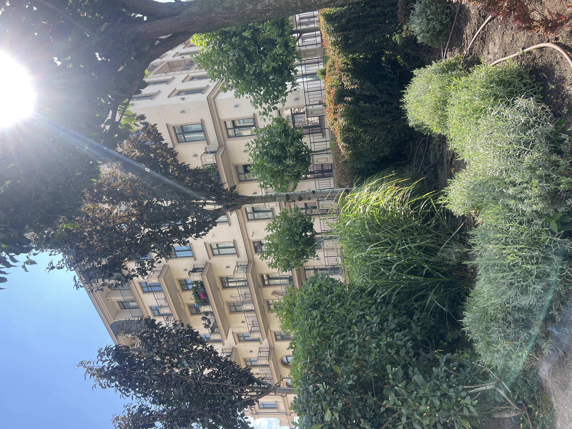 The image depicts a modern apartment building with a beige facade, multiple balconies, and several trees and shrubs in the foreground. The building appears to be several stories high and has a clean, contemporary design. The trees in the foreground are a mix of deciduous and evergreen varieties, with dense foliage. The shrubs are well-maintained and form a neat border along the building's perimeter. The scene is bathed in sunlight, with lens flare visible, suggesting a bright, clear day. The location is identified as Fontanka, Ukraine, and the overall impression is one of urban living with a touch of nature. There are no people or animals visible in the image.