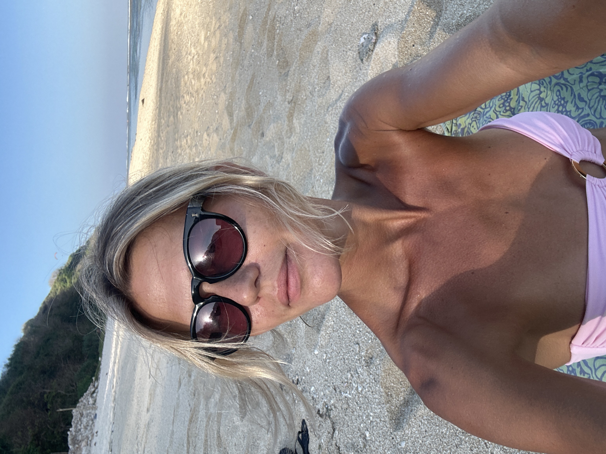 A woman with blonde hair and sunglasses is taking a selfie on a beach. She is wearing a pink bikini top. The beach is sandy with some rocks and vegetation in the background. The ocean is visible in the distance. The sky is clear and blue. The image appears to have been taken in the city of Kuta Selatan, Indonesia, based on the visual cues such as the type of beach, vegetation, and general atmosphere.