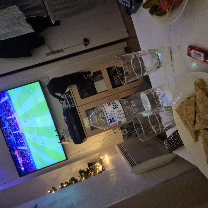 A television displays a soccer game. A black and white cat sits on a cabinet, and Christmas lights are visible. A table is next to the cabinet. It has two glasses, a water bottle, and a plate of crackers. There is also a plate of pickles and some kind of red sausage.