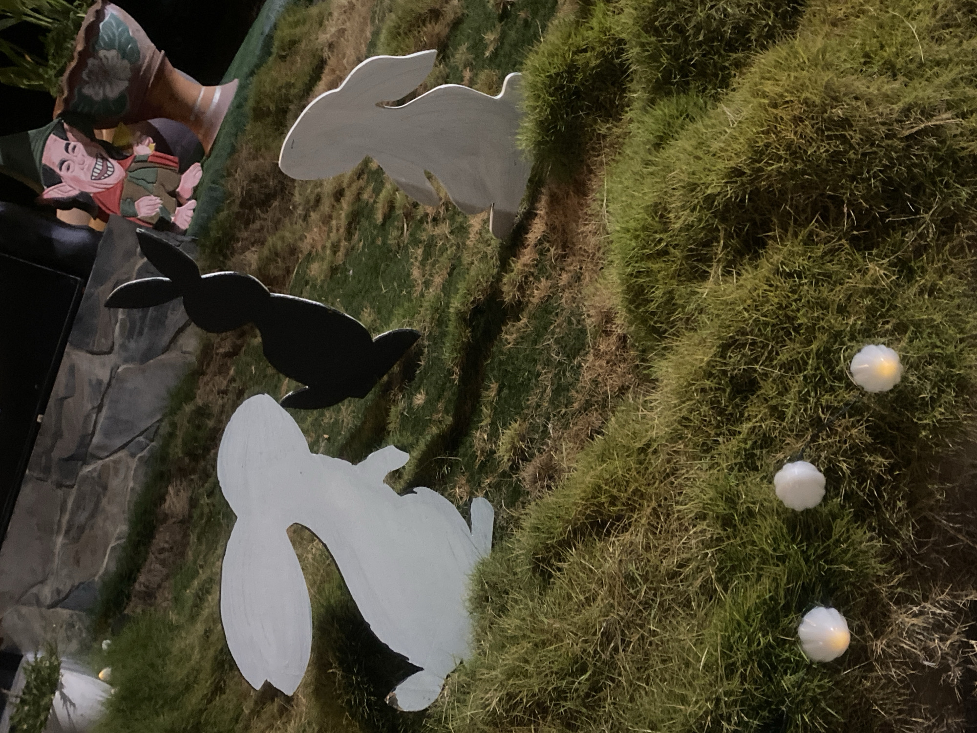 The scene depicts an outdoor Easter decoration in Baan Don Sai, Thailand. Three rabbit silhouettes – two white and one black – are positioned on a grassy area. Adjacent to the rabbits is a decorative gnome figurine emerging from a stylized flowerpot. Three small, round, glowing objects are scattered amongst the grass. A stone-like background is visible to the left.