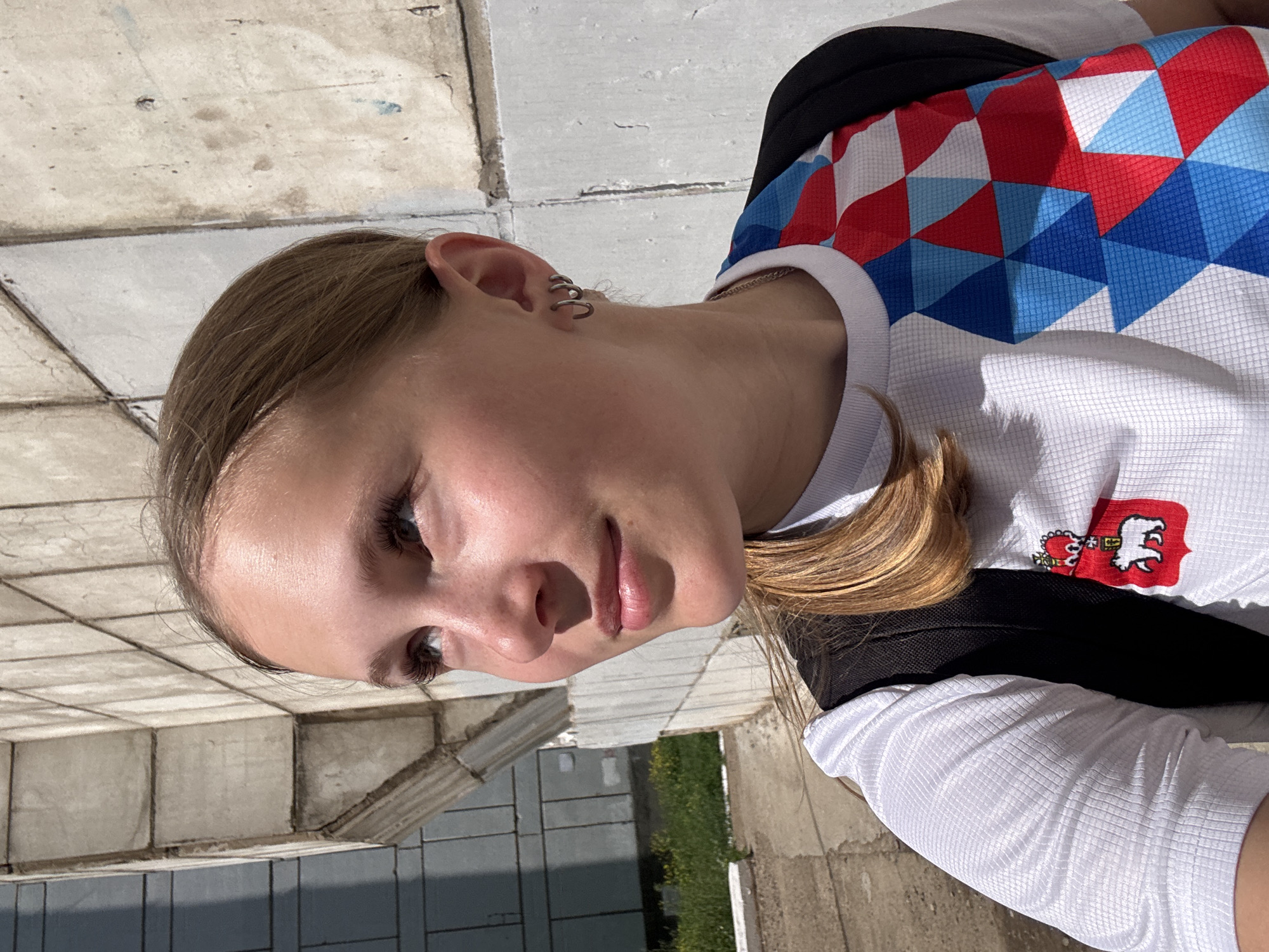 In a medium-wide, sunlit, eye-level shot, a young woman with light brown hair pulled back is pictured from the chest up, looking to her left. She is positioned in front of a textured, light-colored concrete building with a visible shadow line suggesting overcast conditions or a partially shaded area. The building's wall is composed of rectangular panels. To the left of the woman, a patch of green grass and some yellow wildflowers are visible, bordering a concrete walkway.
The woman wears a white t-shirt with a pattern of blue, red, and white geometric shapes on the right side of her chest. A black backpack is visible over her shoulders. On the left side of her chest, there is a red emblem with a white bear and a crown above it, resembling a coat of arms. She has on small hoop earrings in her left ear. The lighting creates bright highlights on her skin, particularly her forehead and nose, and casts shadows on the building, indicating the sun is high in the sky. The overall environment suggests an outdoor urban setting, likely in a public space or near a building. The presence of the emblem on her shirt may indicate affiliation with a specific region or organization within Chaikovsky, Russia, possibly related to sports or local identity.