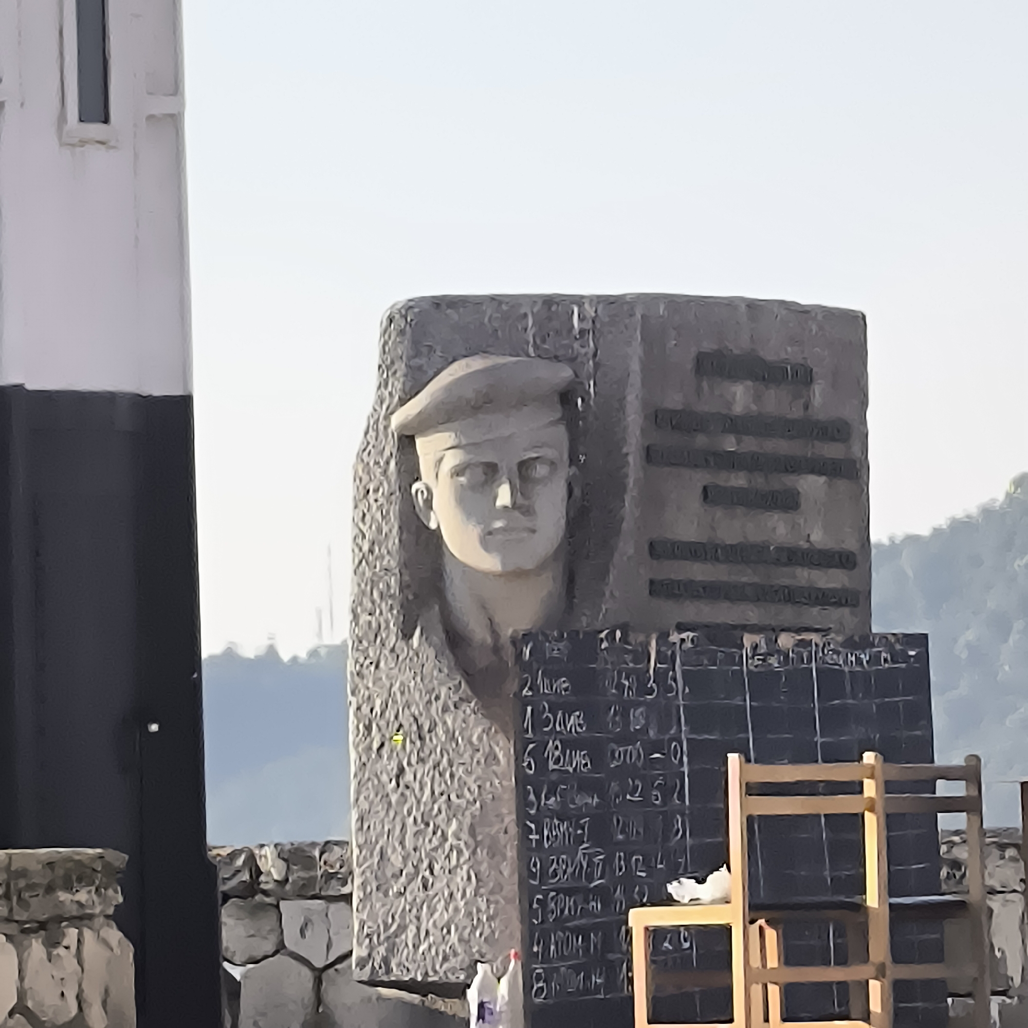 The image shows a monument featuring a bas-relief portrait of a man in a beret, carved into a stone slab. To the right of the monument is a blackboard with chalk writing, appearing to be a list of names and numbers. In front of the blackboard are two wooden chairs. A tall, dark and white structure, possibly a lighthouse, is visible on the left side of the frame. The background shows a hazy sky and distant hills. The monument is located in Varna, Bulgaria, specifically at the Naval Museum, which houses historical artifacts and exhibits related to Bulgarian maritime history.