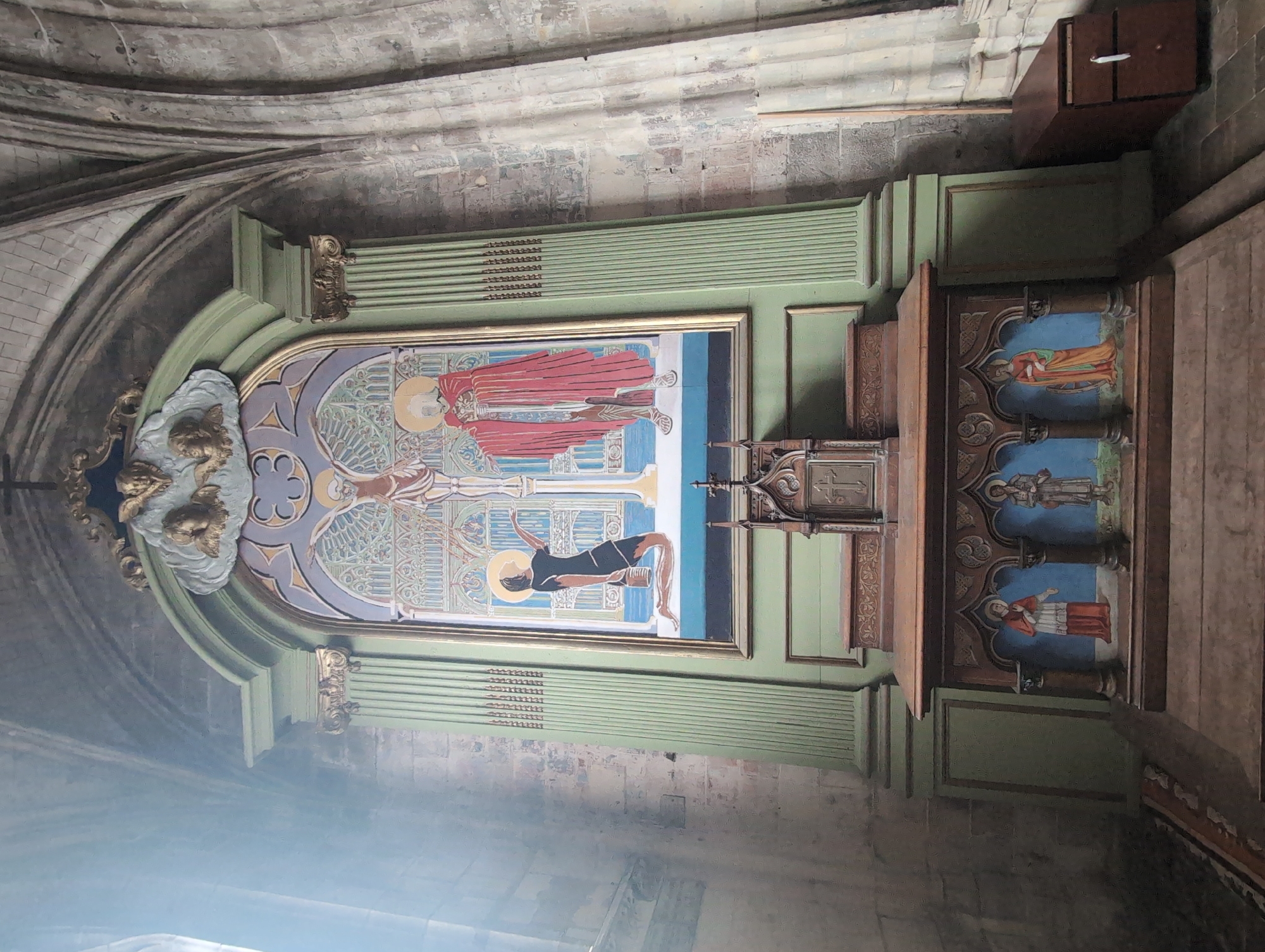 An altar piece is centrally located in a dimly lit, stone-walled interior, likely a church in Le Tréport, France. The altar piece is a tall, multi-layered structure with a prominent painted panel at the top depicting the crucifixion of Jesus Christ. To the left of the cross, a kneeling figure in a black garment holds a bucket, reaching towards the cross. To the right, a figure in a red robe stands with arms outstretched. Above the painting are gold cherubim figures. Below this main panel, a smaller wooden structure houses three painted figures in arches. These figures are dressed in religious garments: one in red, one in blue and white, and another in orange. The entire altar piece is framed by green painted pillars. To the right of the altar piece, a dark wooden chest with drawers sits against the stone wall. The environment is characterized by aged stone architecture, with arched ceilings and walls showing signs of wear.