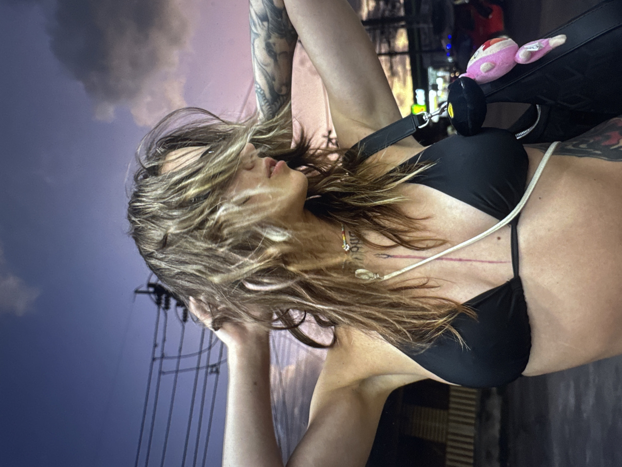 A young woman with long, tousled brown hair and tattoos on her arms is seen from the chest up, posing outdoors in what appears to be a city environment at dusk. She is wearing a black bikini top and has her hands behind her head, looking to her right with her face partially obscured by her hair. A necklace with colorful beads is visible around her neck, and a thin strap from a bag crosses her chest. The bag itself is black with a plush pink toy attached to it. In the background, power lines and a dusky sky with clouds are visible, suggesting an urban setting at twilight. There are no indications that this scene is located in Moscow, Russia.