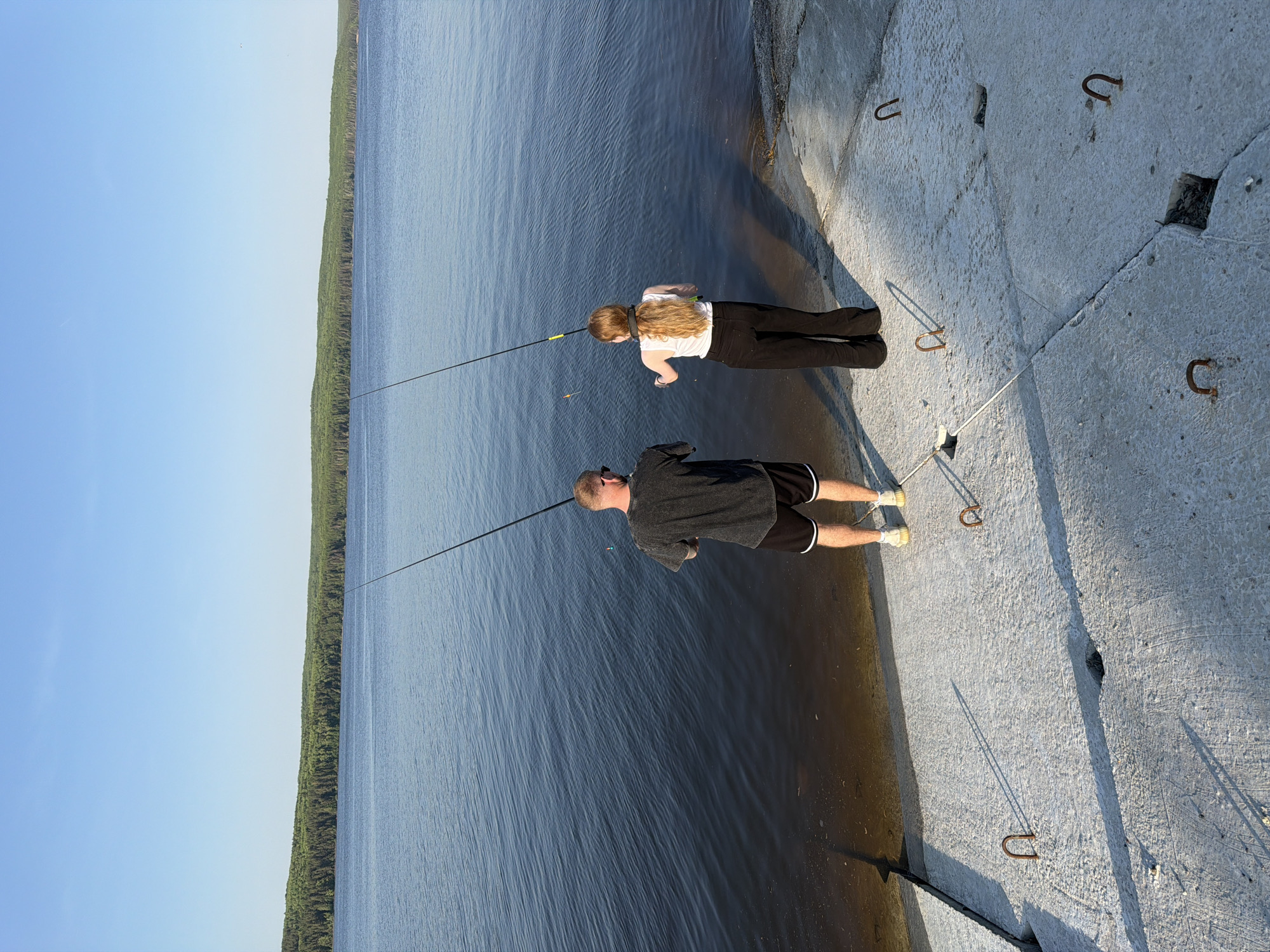 Two people are fishing on a concrete pier alongside a large body of water. A man with a shaved head wearing a dark t-shirt and black shorts stands closest to the viewer. Further down the pier, a woman with long blonde hair is wearing a white shirt and dark pants. Both are holding fishing rods with lines cast into the water. A dense forest lines the opposite bank of the water. The sky is clear and blue. The concrete pier has several metal U-shaped anchors set into it. The location is Chaikovsky, Russia.