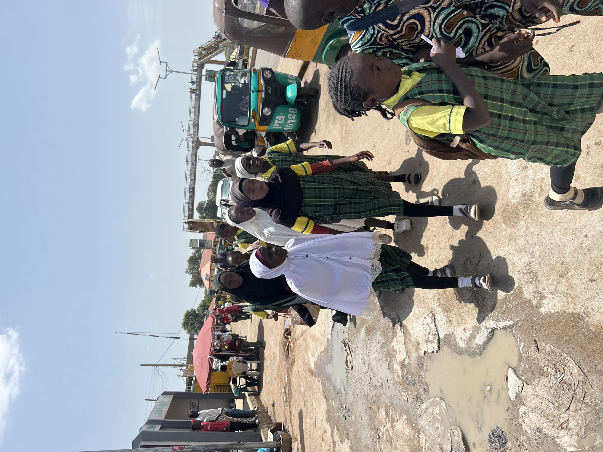 In Jos, Nigeria, on a bright, sunny day, a group of young girls, some in school uniforms consisting of green plaid dresses over yellow shirts, are gathered on a dusty, uneven road. One girl in the foreground wears a white hijab and dress, and another wears a black hijab and dress. They appear to be interacting with each other, with some looking towards the camera.
Behind them, a teal and yellow motorized rickshaw, commonly known as a 'Keke Napep', is parked. The rickshaw has license plates visible, including "PTA- 0128". Several other vehicles, including what appears to be a white van, are also present in the background, suggesting a busy street or thoroughfare.
The environment is characteristic of a developing urban area, with bare earth, cracked pavement, and some puddles of murky water visible on the ground. In the distance, there are buildings, trees, and overhead structures, possibly part of a market or transport hub. A red umbrella is also visible, possibly belonging to a street vendor. The sky is clear blue with a few scattered white clouds. The overall scene captures a slice of daily life in Jos.