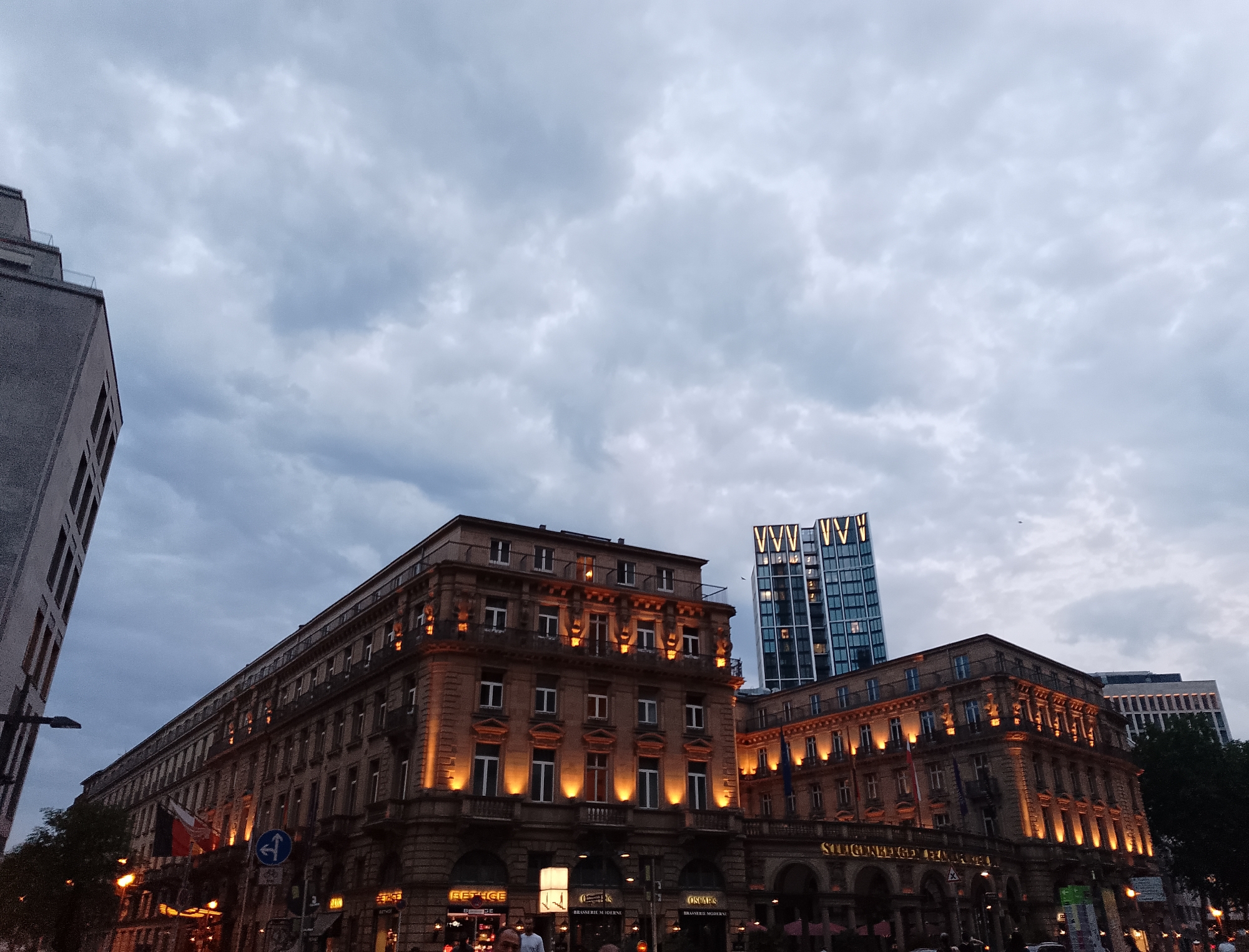 The photograph captures a street scene in a city, possibly in Europe, featuring a cluster of buildings under a cloudy sky. In the foreground, a grand, older building with ornate architectural details and lit windows spans across the frame. The building is illuminated with warm, golden lights highlighting its facade. To the left, a portion of a modern, gray, high-rise building is visible, angled towards the sky. In the background, a slender, contemporary skyscraper with a distinctive "W" shaped facade is prominently displayed, also illuminated with lights. Another modern building with horizontal white window bands stands to the right of the skyscraper. The sky is overcast with a mix of gray and white clouds, suggesting it might be dusk or an overcast day. Several streetlights and signs are visible at street level, indicating an urban environment. Flags are visible on the facade of the older building. The scene is devoid of any clear human activity, with only a few blurry figures visible near the street level.