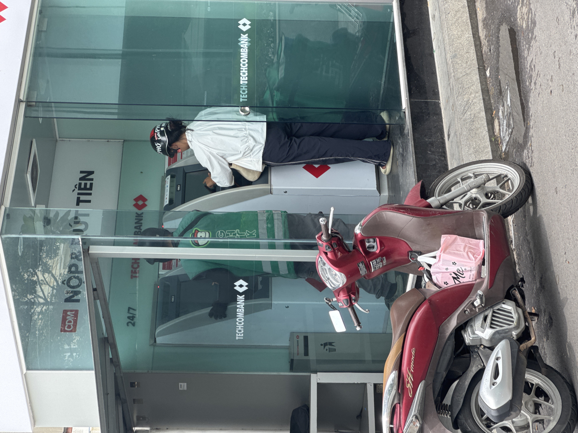 A woman in a white jacket, dark pants, and a helmet is using a Techcombank ATM outside a bank in Nha Trang, Vietnam. She is holding a bag. A red motorcycle is parked in front of the ATM. The bank has a glass exterior with the Techcombank logo visible.
