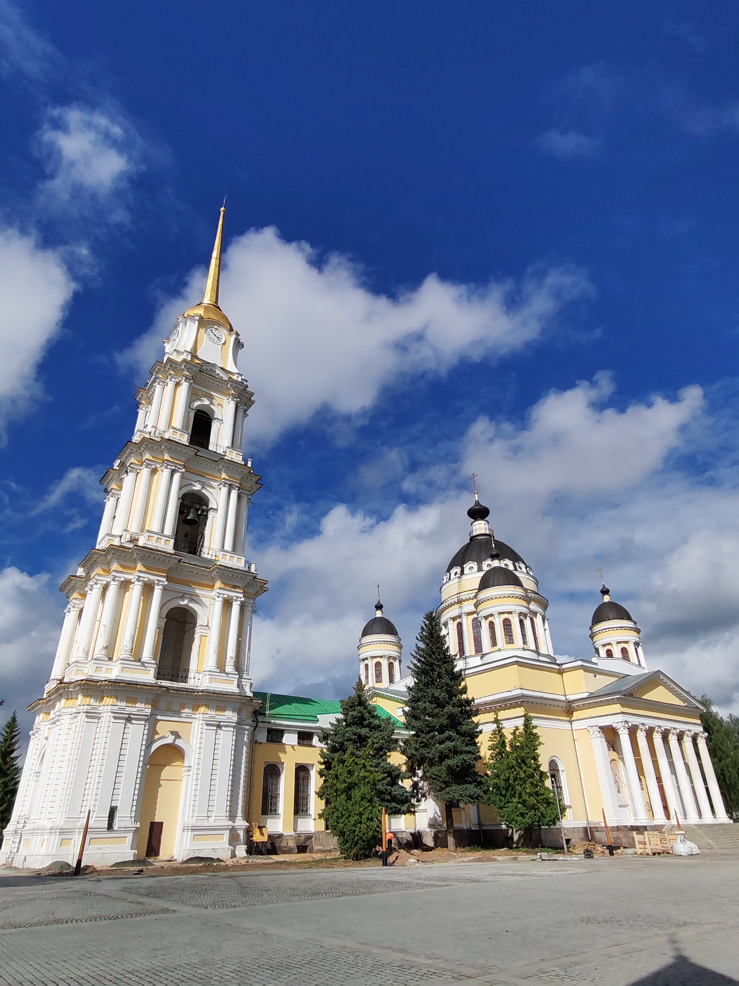 The image shows a large, yellow church with a tall, white bell tower in front of it. The church has several domes, and the bell tower has a gold spire. The sky is blue with some white clouds. There are some trees and a paved courtyard in front of the church. The scene appears to be in the city of Bol'shie Mkhi, Russia. The image captures a clear, sunny day.