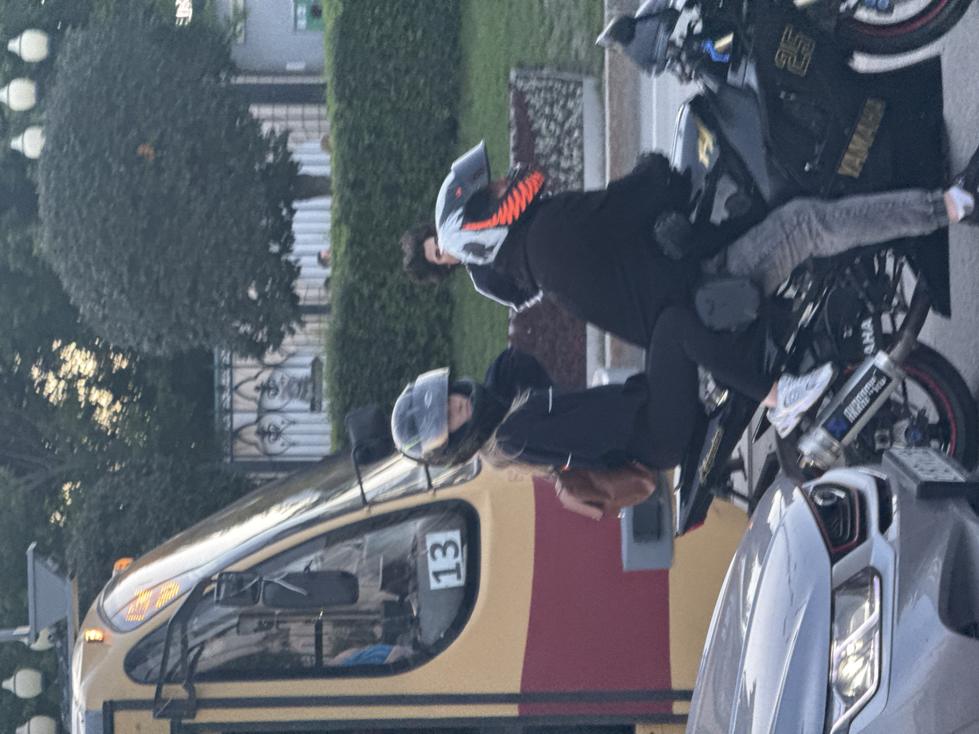The image shows a street scene in Yekaterinburg, Russia, with a yellow and red tram on the left. In front of the tram, two individuals are seated on a black motorcycle. The person in front is wearing a black helmet with green accents and a black jacket, and appears to be female based on hair color. The person behind is wearing a black helmet with orange and white accents and a black jacket, and appears to be male. Both are wearing casual clothing. The motorcycle has "YAMAHA" and "25" visible on its side. To the left of the motorcycle, the front of a silver car is visible. The background features trees, a green hedge, a decorative fence, and lampposts. The overall lighting suggests it is daytime, possibly late afternoon or early evening, with some sunlight filtering through the trees.
