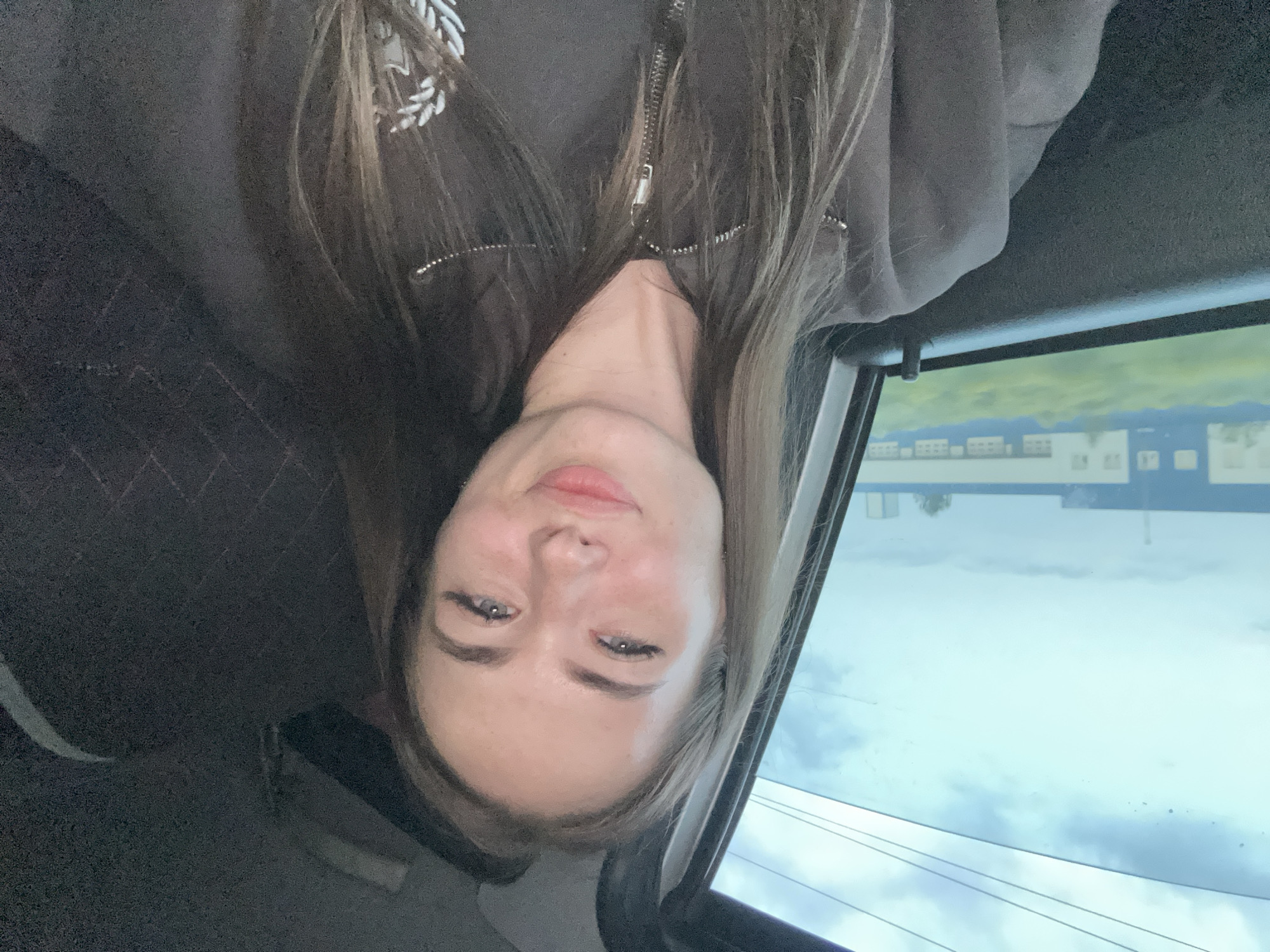 A young woman with long brown hair and blue eyes is captured in a selfie taken from inside a vehicle. She is wearing a dark gray zip-up jacket. Her head is tilted slightly, and she is smiling with her lips parted. In the background, through the car window, a building complex with a blue roof and white walls is visible, surrounded by green foliage. The sky above is cloudy and overcast. The interior of the car, including the dashboard and a dark textured seat, is also visible.
