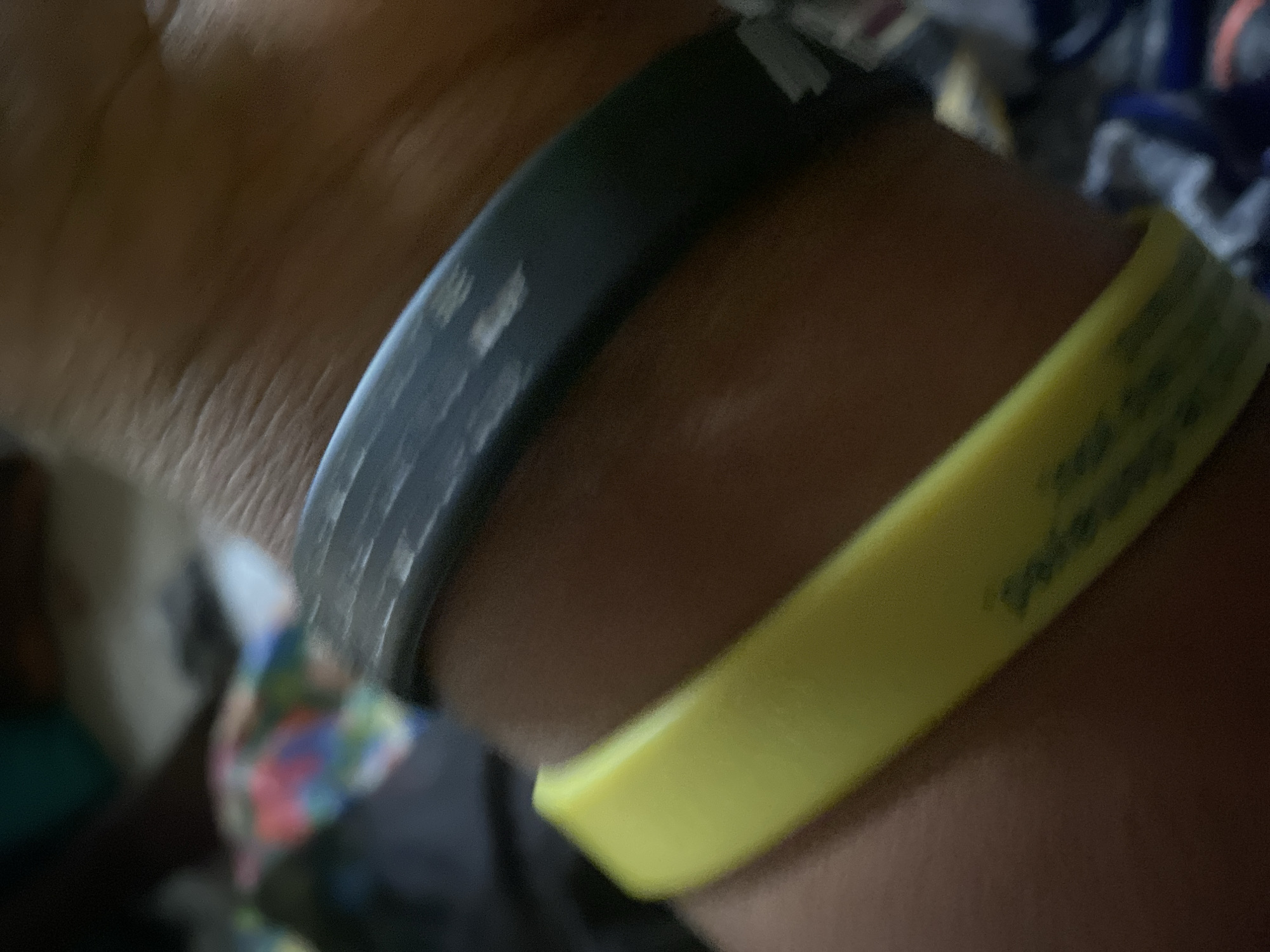 The image shows a close-up of a person's wrist wearing two silicone wristbands. The upper wristband is black and appears worn, with scuff marks and some text partially visible. The lower wristband is bright yellow and also shows some dark text or markings. The skin texture is visible, suggesting a close-up shot. The background is blurred and appears to be indoors, with some colorful fabric or clothing visible. There is no discernible connection to Jauro Yinu, Nigeria, as the image is a close-up and lacks any contextual environmental or geographical clues.
