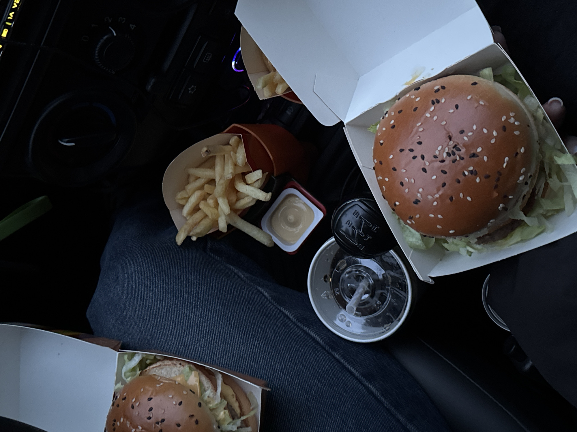 The image depicts a McDonald's meal spread across a person's lap in what appears to be the front seat of a car. The meal consists of two burgers in open cardboard containers, two servings of french fries, a sauce container, and a soft drink with a lid and straw. The person is wearing jeans. The car's interior, including a portion of the dashboard with knobs, is visible. There is no evidence of the location being Perm, Russia in this image.
