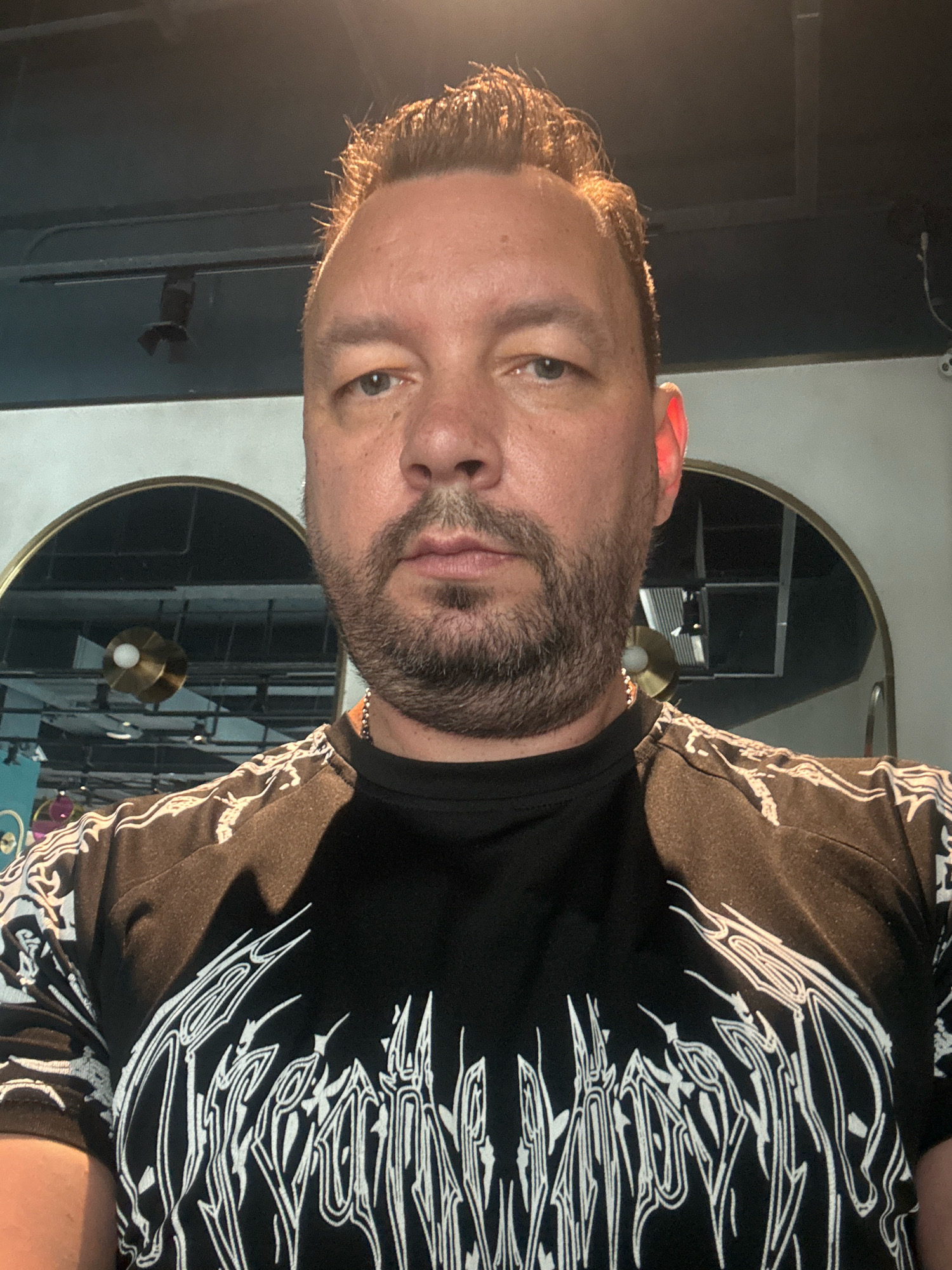A man is seen in a close-up, front-facing selfie. He has short, styled brown hair with highlights, a beard and stubble, and is wearing a black t-shirt with a white, abstract graphic design on it. The background shows interior architectural elements, including a ceiling, arched mirrors, and lighting fixtures. The lighting appears to be artificial, with a reddish hue on the man's right ear. The image appears to be taken indoors. No specific location in Nanhai District, China can be determined from the image.