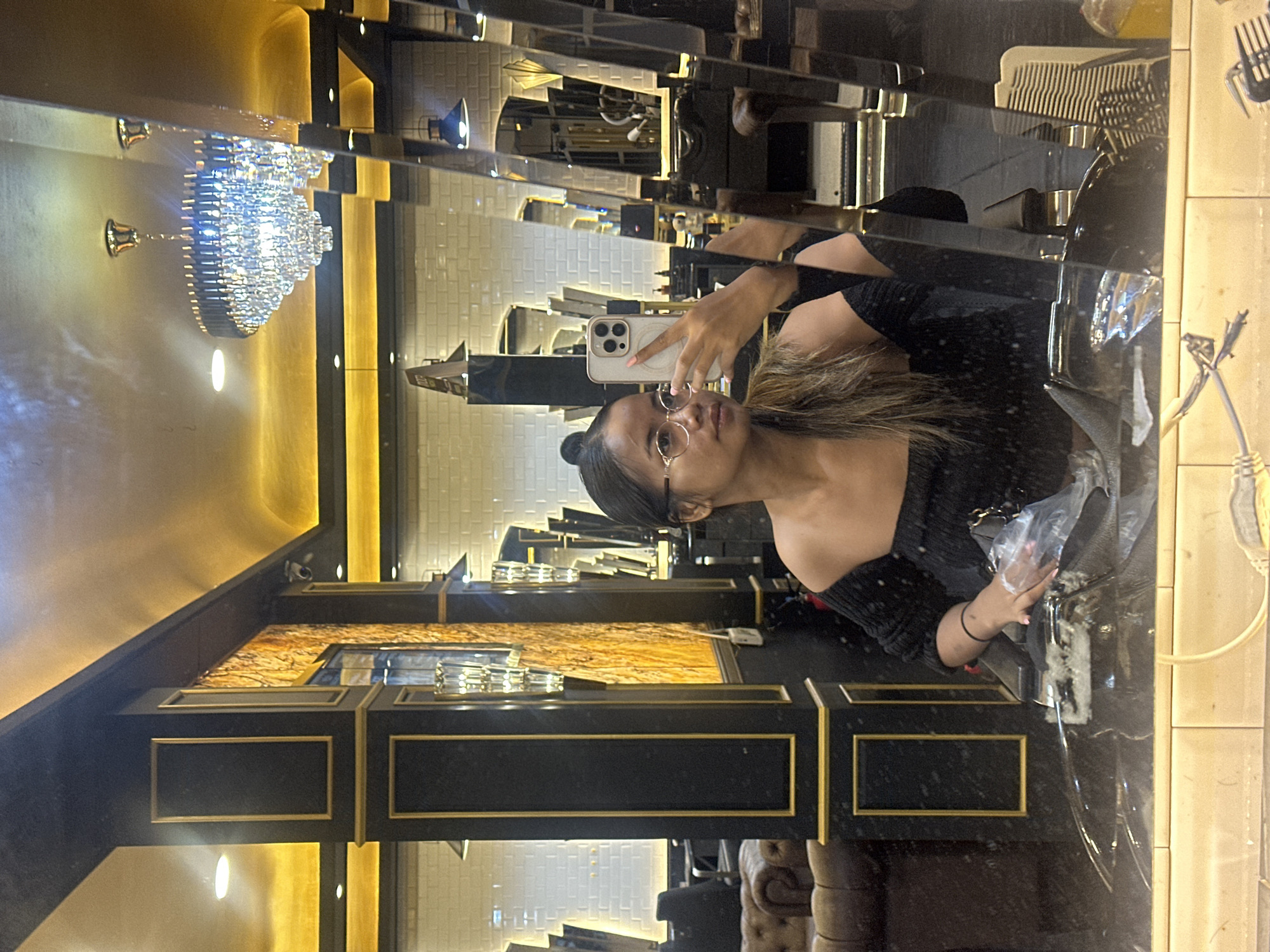 A young woman captures a mirror selfie in a luxurious indoor setting, likely a high-end salon or boutique in North Jakarta, Indonesia. The image is rotated sideways, showing the woman's reflection upright when viewed correctly.
The main subject is a woman with dark hair pulled back, wearing glasses with thin frames and a black off-the-shoulder top. She holds a light-colored smartphone with multiple cameras up to take the photo, reflecting her arm and the device. Her expression is neutral, focused on the selfie. In her other hand, she holds a clear plastic bag.
The background is opulent, dominated by a scheme of black, gold, and warm lighting. A large, multi-tiered crystal chandelier hangs prominently on the left, casting sparkling light. The space features black decorative pillars or columns with intricate gold trim, reflecting multiple angles of the room. These structures and surrounding walls are highly reflective, creating a complex array of reflections. Behind the reflections, a wall adorned with white rectangular tiles, possibly subway tiles, is visible. On a surface to the right, a white comb or brush and other styling tools can be seen, reinforcing the impression of a salon. Part of a dark, tufted piece of furniture, possibly a sofa or chair, is visible in the lower right.
The overall atmosphere is one of sophistication and elegance, suggesting an evening or indoor setting where artificial light provides the primary illumination. No discernible text is visible.