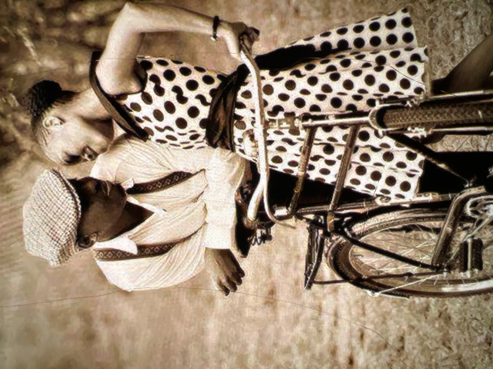 In a sepia-toned, artistic rendering of a scene possibly set in Lagos, Nigeria, a Black man and woman are depicted in a romantic embrace beside a bicycle. The man, wearing a white button-down shirt with suspenders and a checkered flat cap, leans towards the woman. She is dressed in a white dress with large black polka dots and a dark sash, her hair styled in braids. They are positioned on what appears to be a dusty or unpaved surface, suggested by the textured ground and sparse foliage in the background, characteristic of an outdoor, possibly rural or less developed urban area within Lagos. The bicycle, a vintage model, sits between them, with the woman's hand resting on the handlebars. The overall mood is one of affection and nostalgia.