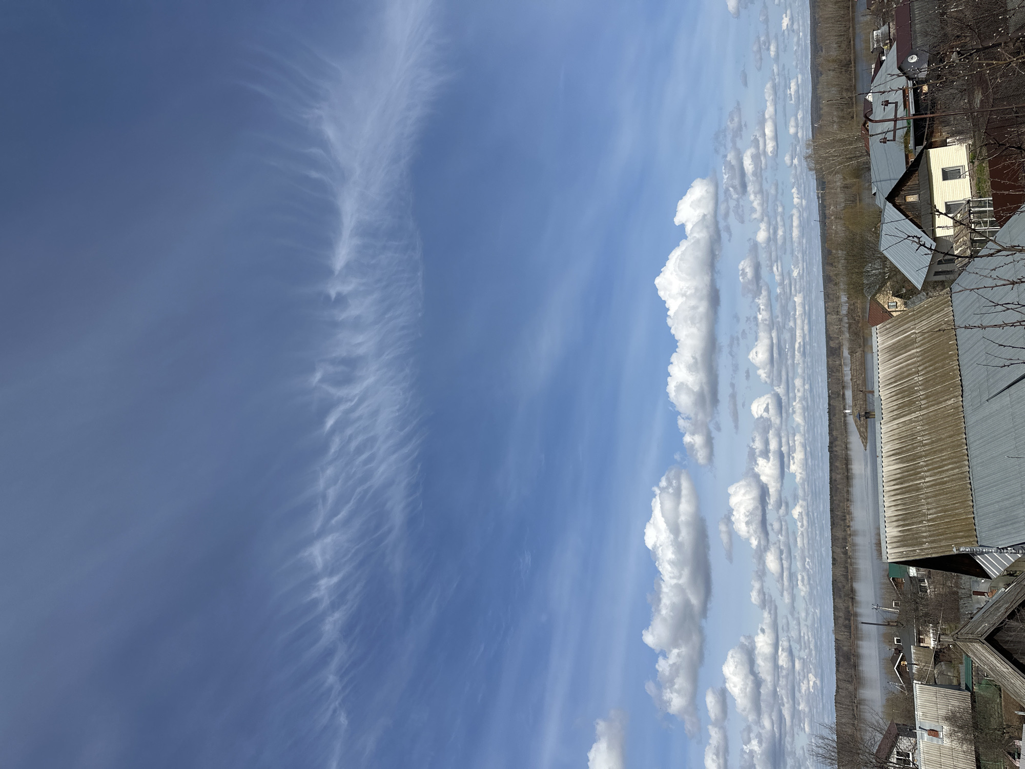 The image shows a view in or near Chaikovsky, Russia, likely taken from an elevated position such as a building or hill. The sky takes up a significant portion of the frame, displaying a mix of cirrus and cumulus clouds. The cirrus clouds are wispy and feather-like, while the cumulus clouds are puffy and white. Below the clouds, there's a landscape featuring a wide body of water, potentially a river or large lake. The water reflects the sky, creating a mirroring effect. The edges of the water are lined with trees, suggesting a wooded area. A line of houses can be seen along the right side of the frame, indicating a residential area. The houses have typical Russian architecture, with pitched roofs and modest designs. The foreground includes parts of buildings, possibly the roof or side of the structure from which the photo was taken, adding depth to the scene. The weather appears to be clear, with good visibility and natural lighting.