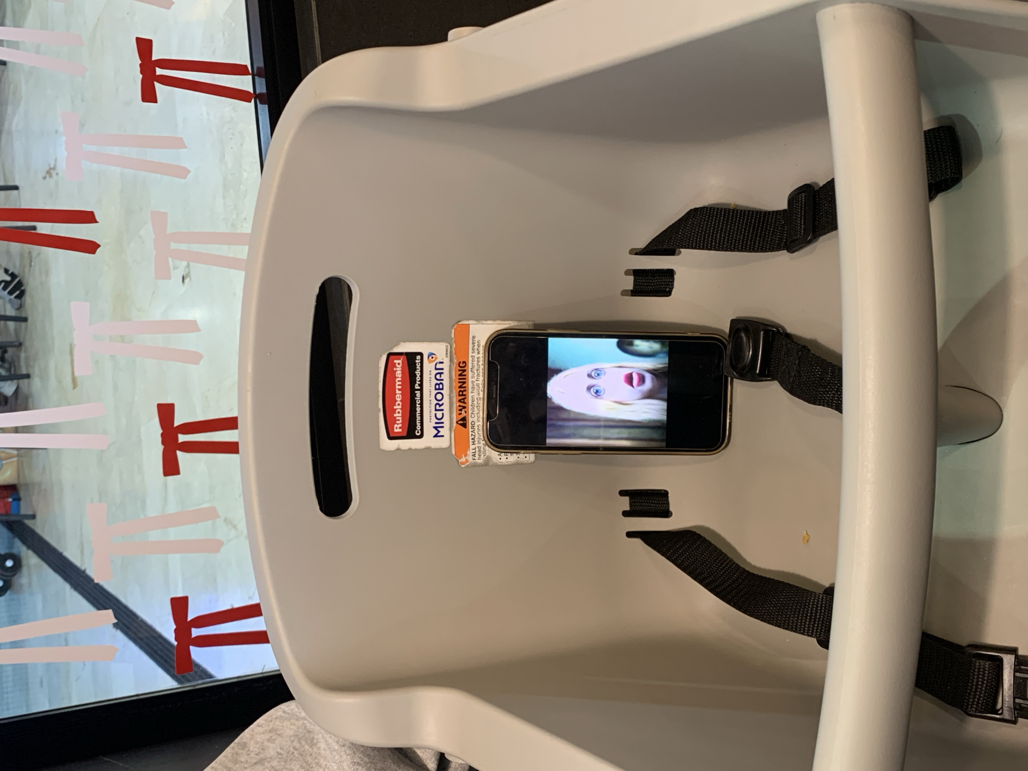 The image shows the inside of a highchair, featuring black straps, a white frame, and several attached warning labels. A smartphone rests inside the highchair, displaying a video of a person with blonde hair and wide eyes. A window in the background is decorated with ribbon-like shapes in white and red. The location is not discernible.