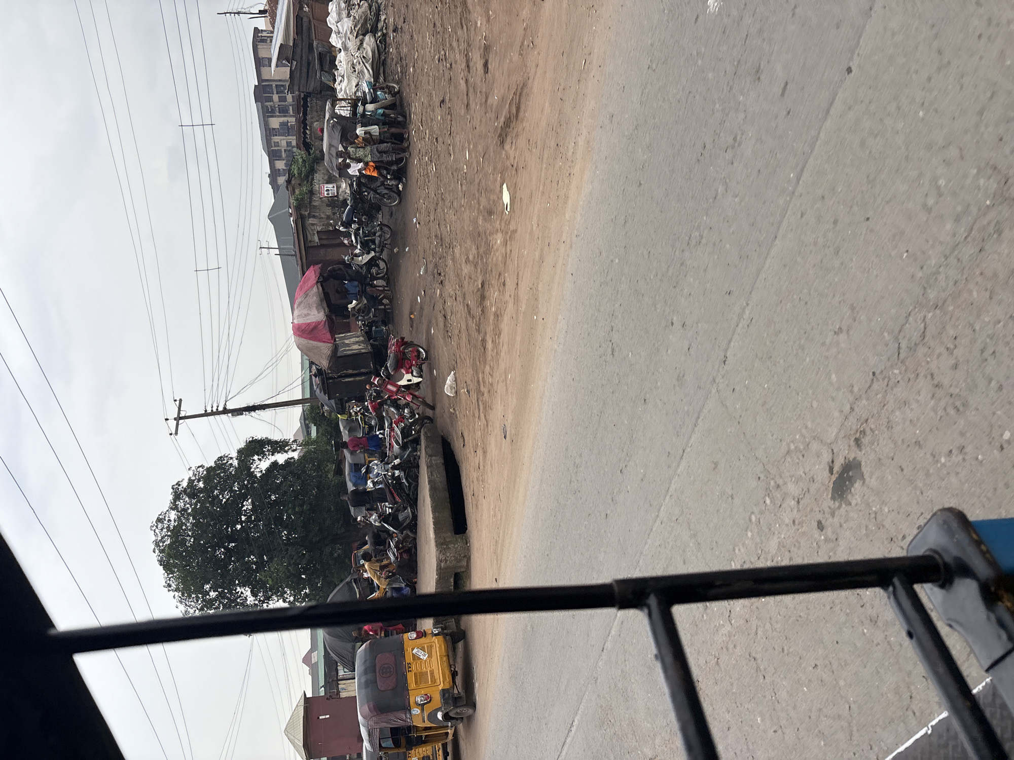 The image depicts a busy street scene in Onitsha, Nigeria, characterized by a lively market or transportation hub. In the foreground, a yellow auto-rickshaw, commonly known as a "Keke Napep," is visible on the left side, suggesting a mode of public transportation. The street itself is paved with asphalt and appears somewhat weathered, with dirt and debris scattered across its surface.
Behind the auto-rickshaw, a collection of motorcycles and other vehicles are parked, indicating a commercial area. Several individuals are present, some appearing to be interacting with the vehicles, possibly as drivers or customers. There are makeshift stalls or shelters with tarps, suggesting a market environment where goods are sold or services are provided. The background reveals a mix of structures, including low-rise buildings and what appear to be more modern apartment blocks further in the distance. Overhead, a network of power lines crisscrosses the sky, typical of urban infrastructure. A large, leafy tree stands prominently in the mid-ground, adding a touch of nature to the otherwise urban setting. The overall atmosphere suggests a bustling, everyday scene in the city.