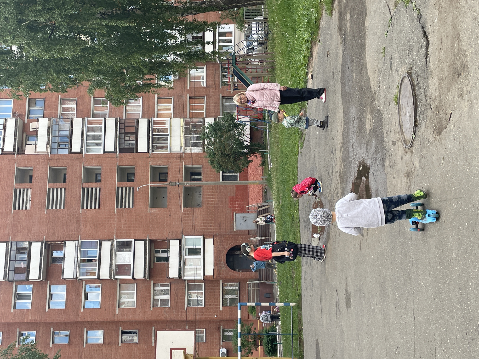 The image depicts an outdoor scene in a residential area of Izhevsk, Russia. In the foreground, a child in a white shirt, camouflage pants, green shoes, and a light-colored hat is lying on a scooter on a gray pavement area. Another child wearing a red jacket is playing nearby. Standing upright is an individual in plaid pants and a red shirt. Beyond them is a grassy area where a woman in a pink jacket is standing with a small child. In the background is a large brick apartment building with numerous windows. There is a playground visible in the background, as well as a basketball hoop and a soccer goal. A large tree is also present near the top of the frame. The ground is wet in places, suggesting recent rain or puddles.