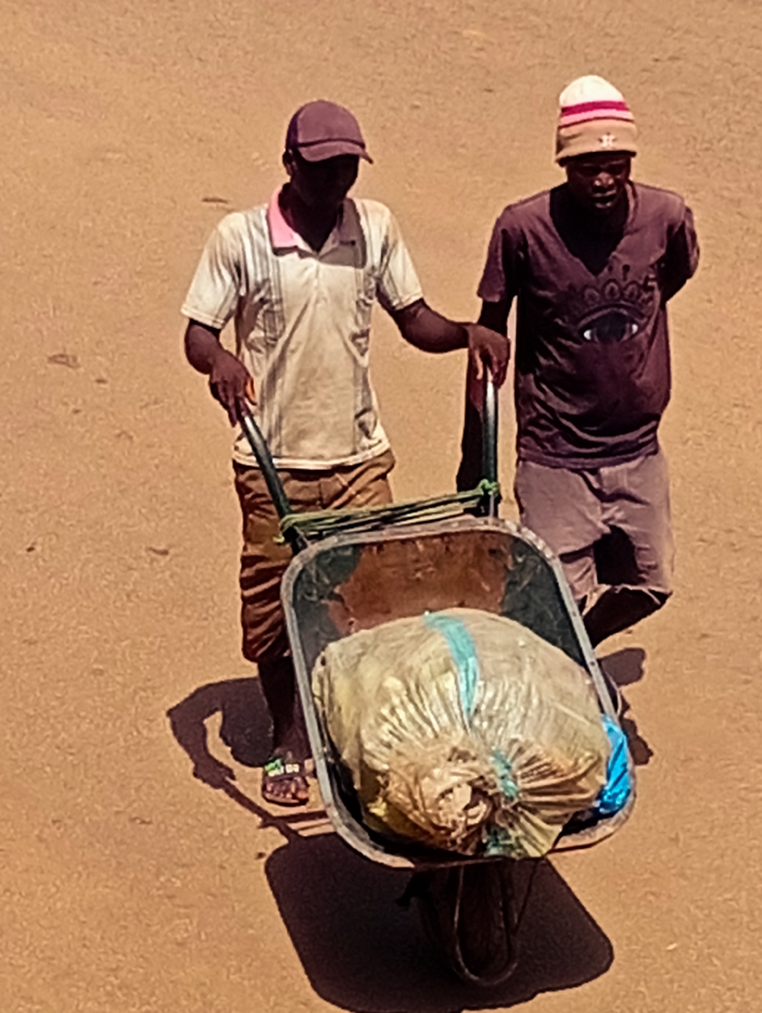 In Bukuru, Nigeria, two men are pushing a wheelbarrow loaded with a large, light-colored bag. The man on the left, wearing a white polo shirt with pink trim and a purple cap, is pushing the wheelbarrow. He is wearing brown shorts and sandals. The man on the right, wearing a dark t-shirt and a beanie with red and white stripes, walks beside the wheelbarrow, his hands behind his back. The ground appears to be a dusty, reddish-brown surface. The lighting suggests it is daytime, with distinct shadows cast by the men and the wheelbarrow.