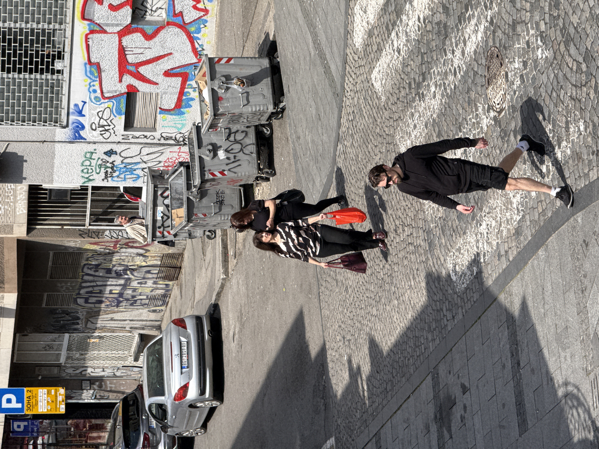 This media file captures an urban street scene in Belgrade, Serbia, on a bright, sunny day. The image is taken from a slightly elevated perspective, looking down on the street and sidewalk.
Three individuals are visible walking on a cobblestone sidewalk on the right side of the frame, casting prominent shadows. The leading person is a man with a beard and sunglasses, dressed in a black hoodie and black shorts, wearing black sneakers with white socks. Behind him, a woman wearing a black and white patterned long-sleeve top and black pants carries a dark red handbag. Partially obscured by her is another woman in black clothing, holding a bright orange bag.
On the left side of the image, an asphalt road features several parked cars, including a silver sedan with the license plate "BG-2889-RE". A yellow and blue parking sign with the text "P", "ZONA 2", and "3OHA 2" (Cyrillic for Zone 2) is clearly visible near the parked vehicles.
The background consists of buildings with walls extensively covered in graffiti, including discernible tags like "KEPA" and "SOMA". Several large, grey waste bins, also adorned with graffiti, are placed along the building front. An older person, possibly a man with grey hair, can be seen looking out from a window opening on one of the buildings.
The strong shadows and bright illumination indicate it is daytime, likely mid-morning or afternoon. No specific emotions are apparent; the people seem to be engaged in everyday activities.