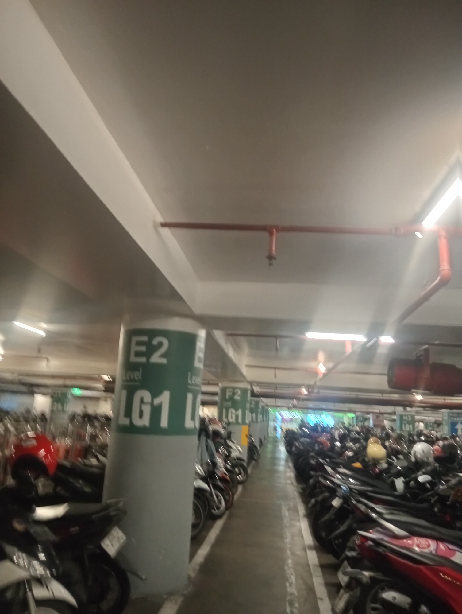 This is an indoor, multi-level parking garage for motorcycles. The scene is dimly lit, with overhead fluorescent lights and the beam of headlights from some vehicles. The garage is filled with rows of parked motorcycles, many of them dark in color. Some of the motorcycles have bright red or white seats or body panels, and one has a yellow helmet visible. There are structural pillars with green and white signage indicating levels and zones, such as "E2 LG1". The floor is concrete with white painted lines marking parking spaces. In the background, there's a bright, colored light installation suggesting a transition or entrance area. There are no people visible in the image. The overall atmosphere is one of a busy, functional urban parking facility.