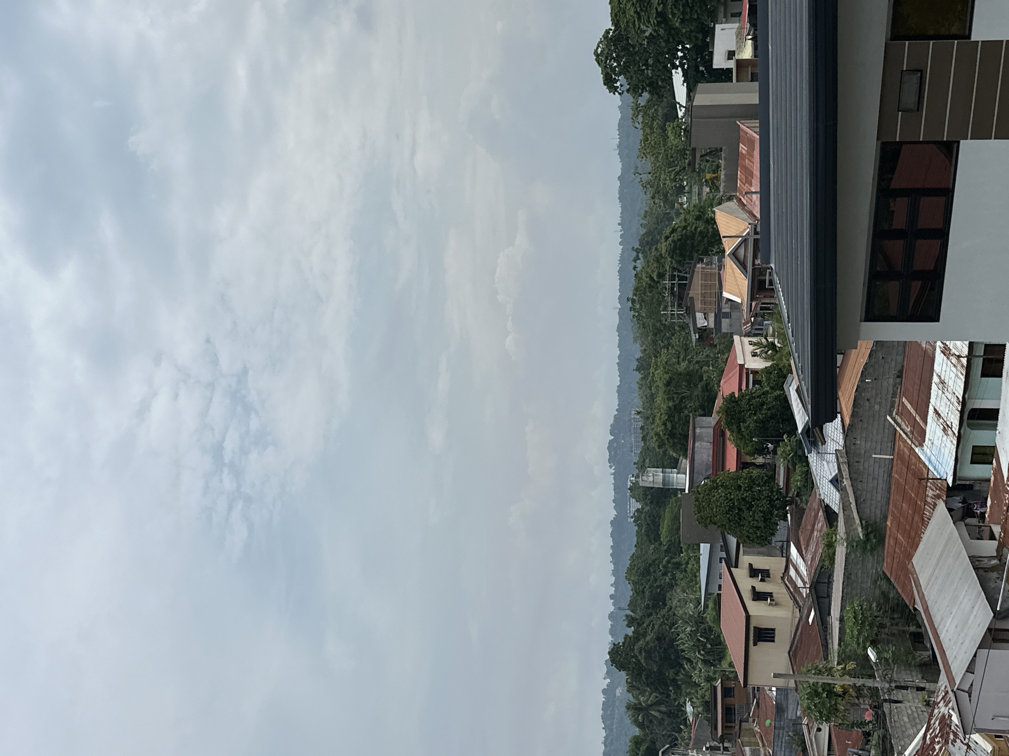 The image depicts a cityscape in Davao City, Philippines, under an overcast sky. Dense vegetation and rolling hills form the backdrop. Houses with varied roofing materials, including corrugated metal and tiles, are clustered together. A tall, white, possibly unfinished building dominates the foreground, showcasing dark window frames and a modern design. Other buildings are visible amidst the foliage, suggesting a mix of residential and commercial structures. No people are visible. The overall scene suggests a densely populated urban area blending into a hilly, green landscape.