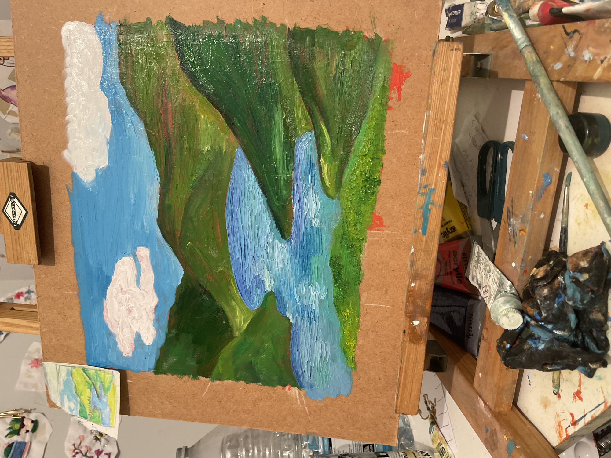 The image shows an oil painting in progress, depicting a mountainous landscape with a river or lake winding through it. The painting is on a brown board, likely particleboard, and is secured to a wooden easel. The painting features broad, expressive brushstrokes. Greens dominate the landscape with blues in the water. There is also an area of white paint that could be snow or clouds. To the right of the canvas is an easel with scattered paint tubes, brushes, and a dirty rag. No people are visible. A small drawing or print is attached to the bottom left of the canvas. The location is not discernable from the image.