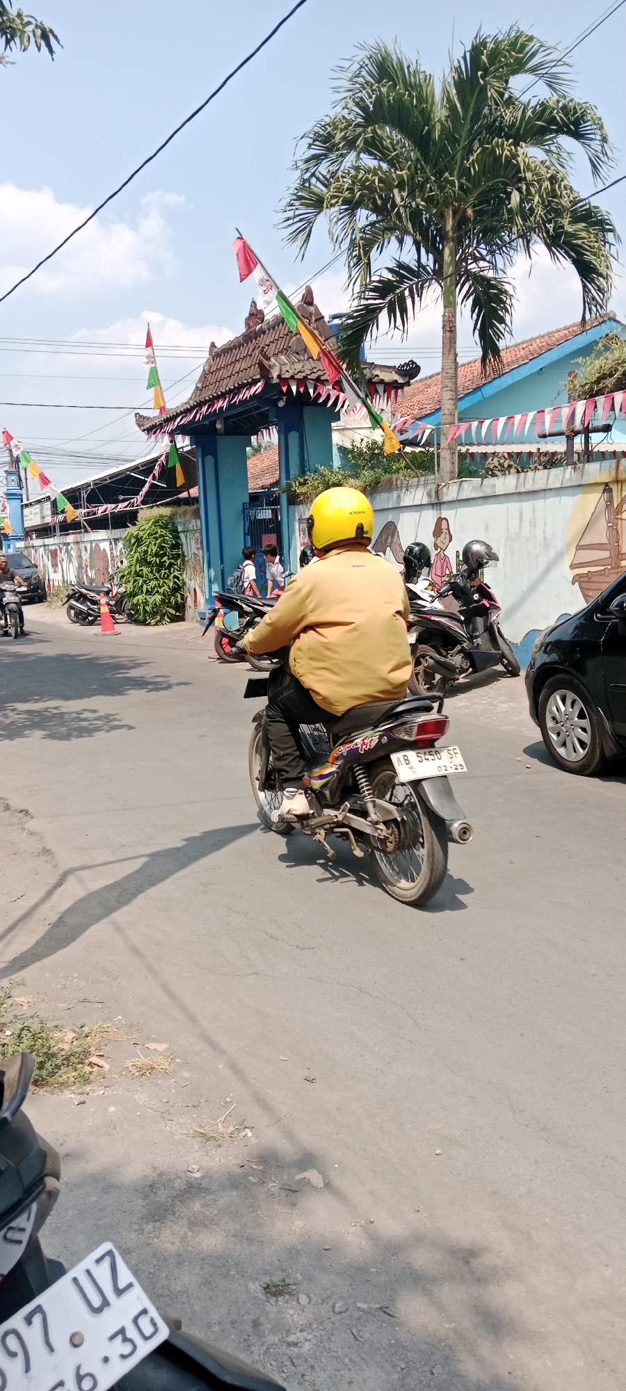 In Purwomartani, Indonesia, a person wearing a yellow helmet and a yellow jacket rides a motorcycle down a street under a clear sky with a palm tree visible. The motorcycle has a license plate that reads "AB 5450 SF 02-29". In the background, a blue building with a traditional roof, adorned with flags, appears to be an educational institution. Other motorcycles, a black car, and several pedestrians are also present on the street. The scene is illuminated by sunlight, casting shadows on the asphalt road.